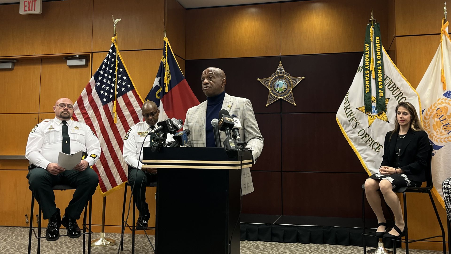 Three men and one woman at a press conference in a sheriff's office. American and North Carolina flags are behind them, along with a sheriff's star emblem on the wall.