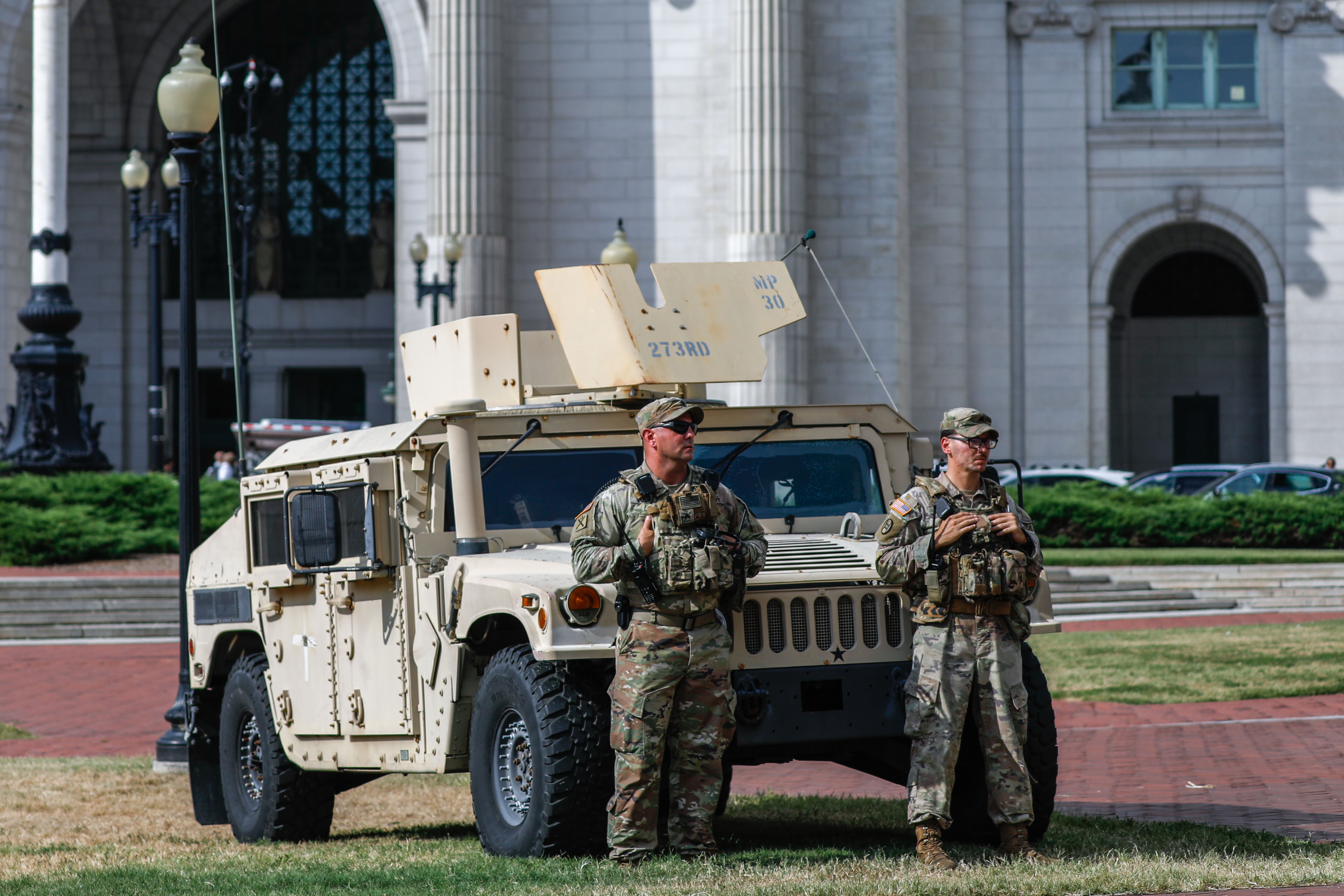 Two soldiers in camouflage gear stand in front of a beige military Humvee outside a large stone government building with arches and windows.