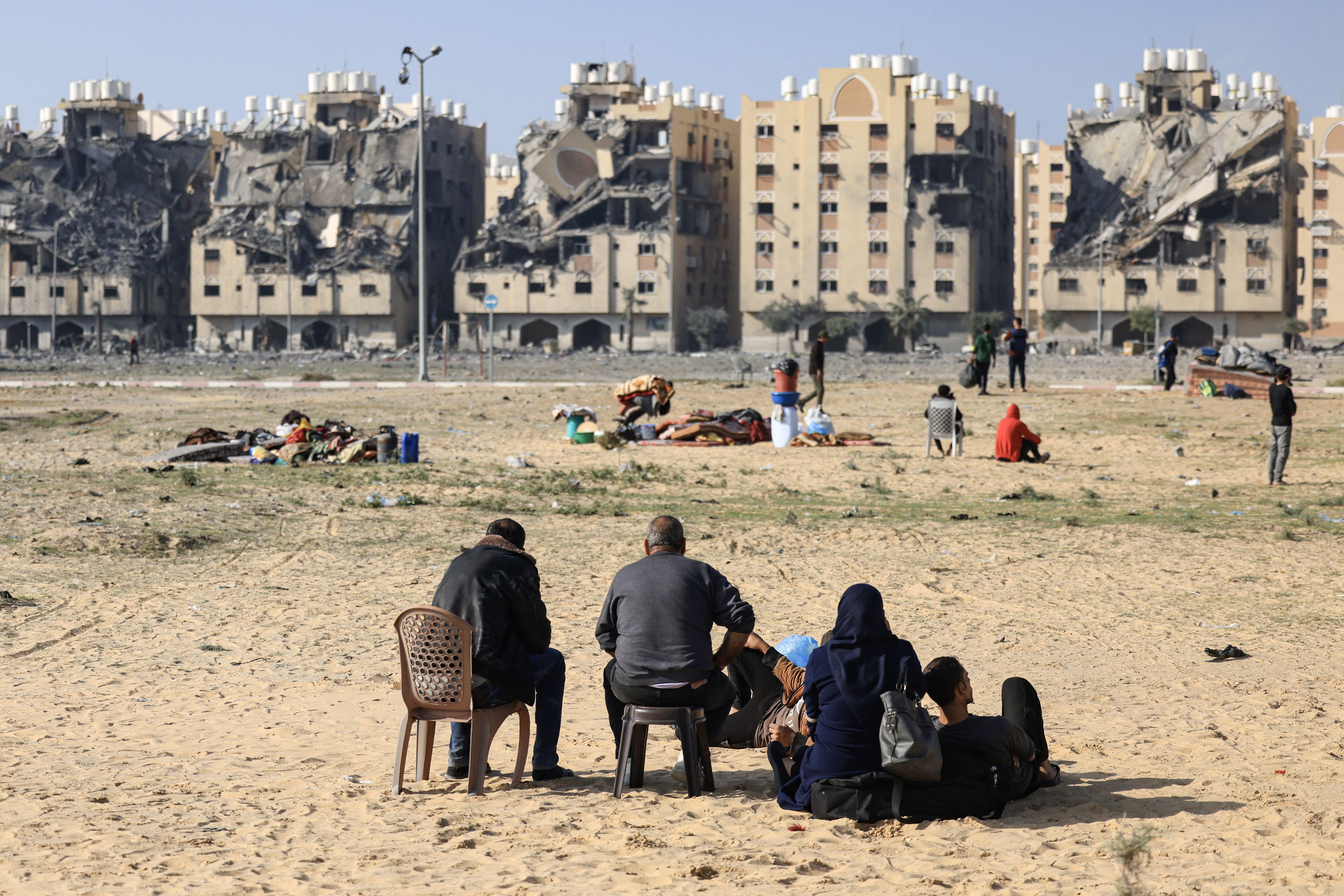 Residents sit in the foreground with their belongings and their backs turned to the camera. In the background are destroyed residential buildings.