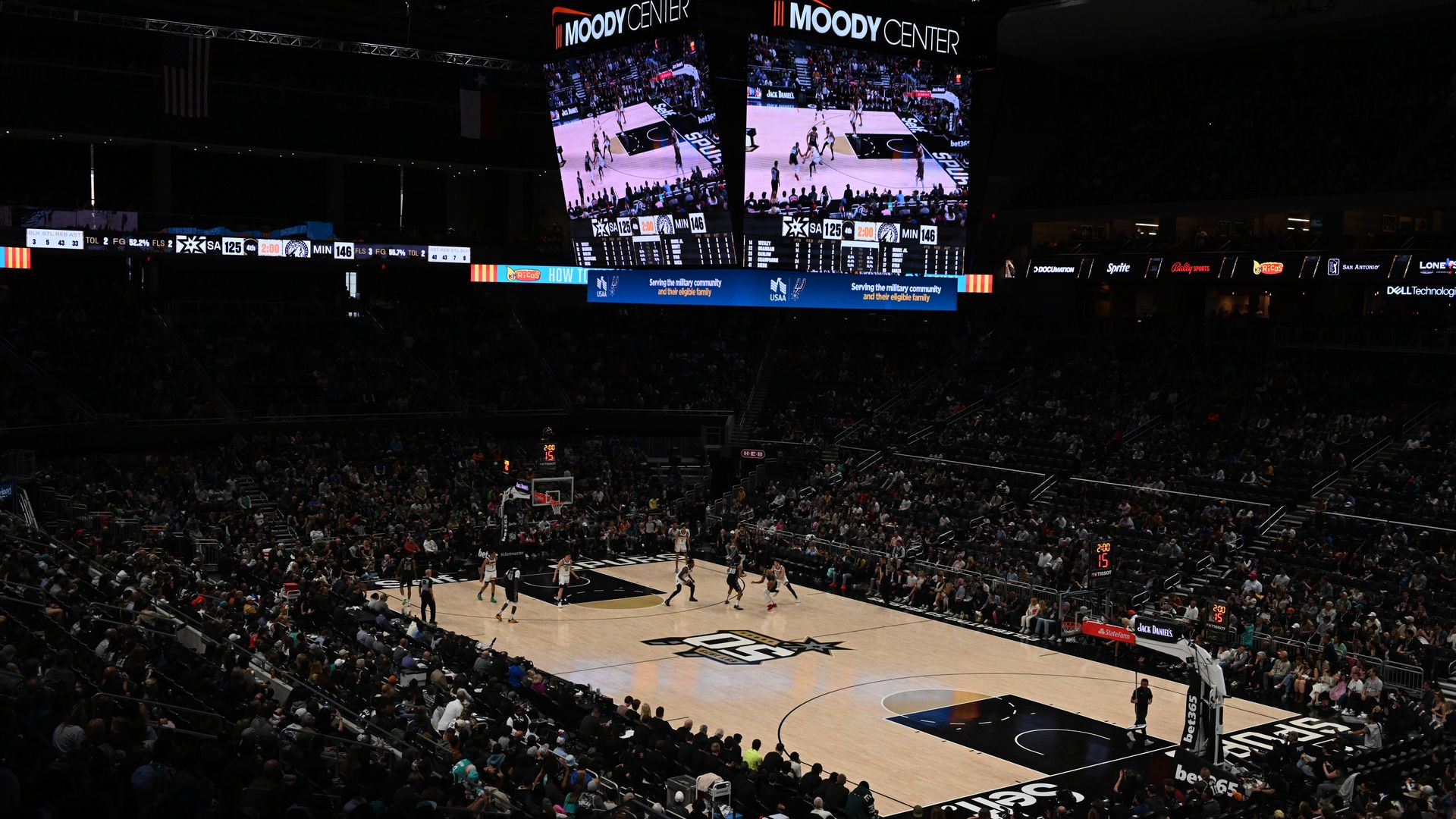 An arena, filled with people, with a Spurs-branded basketball court in the middle. 