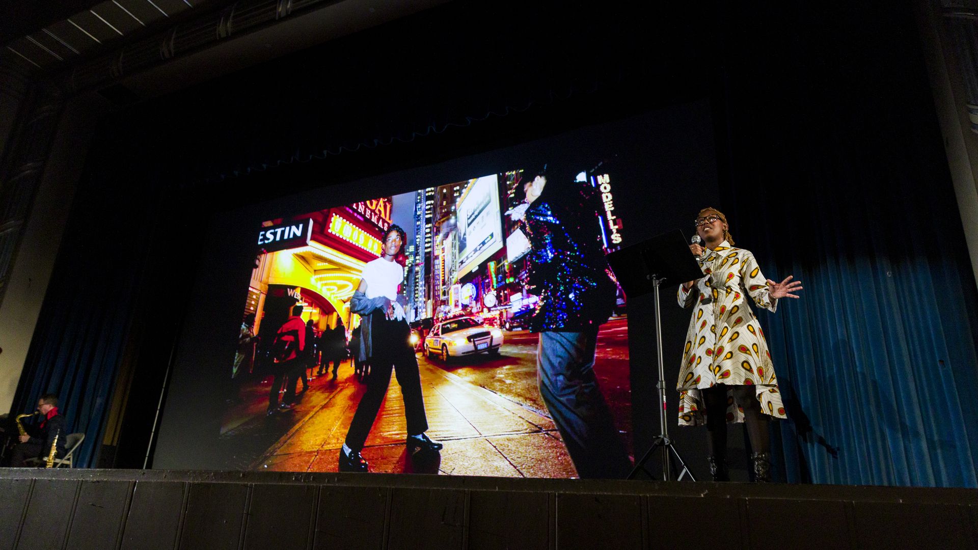 A person on stage speaking with a photo displayed by a projector in the background.