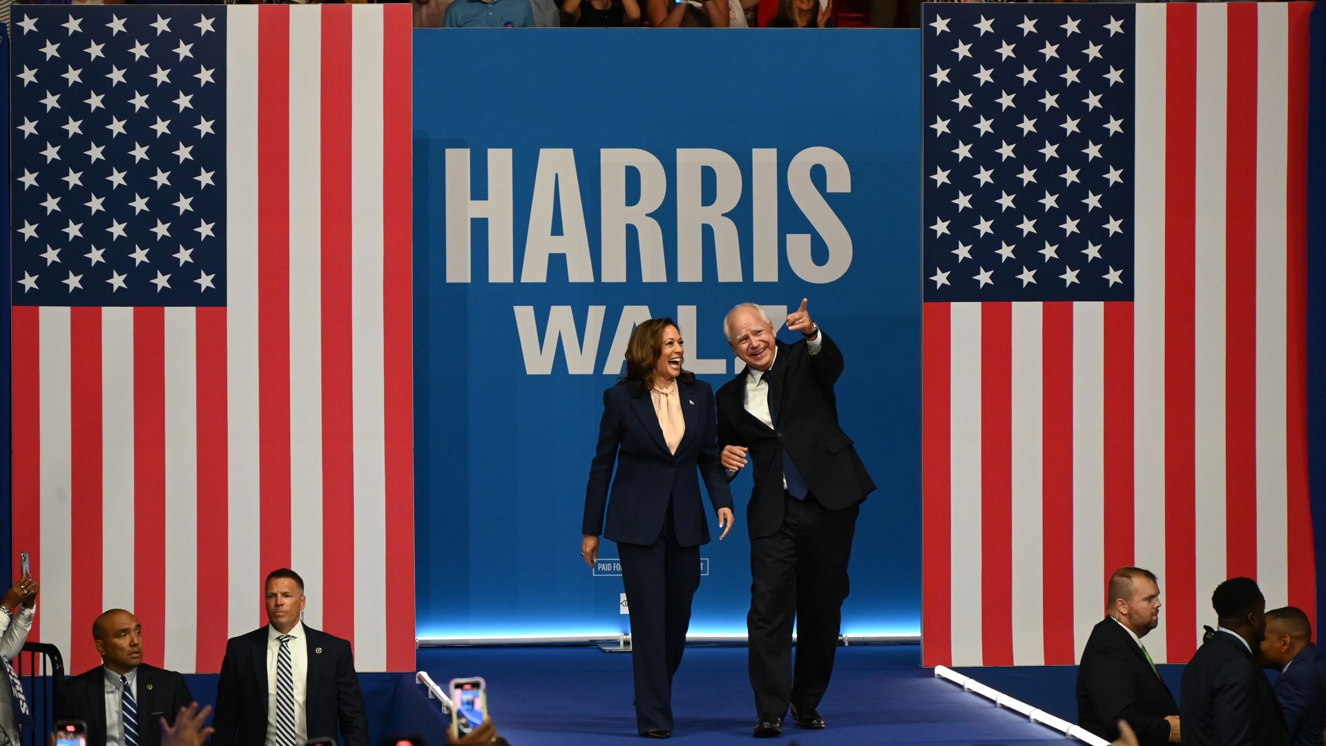 Kamala Harris and Tim Walz walk down a stage with two American flags in the background as well as a sign that says 'HARRIS WALZ.' Hands of people are seen holding phones up taking photo or videos. 