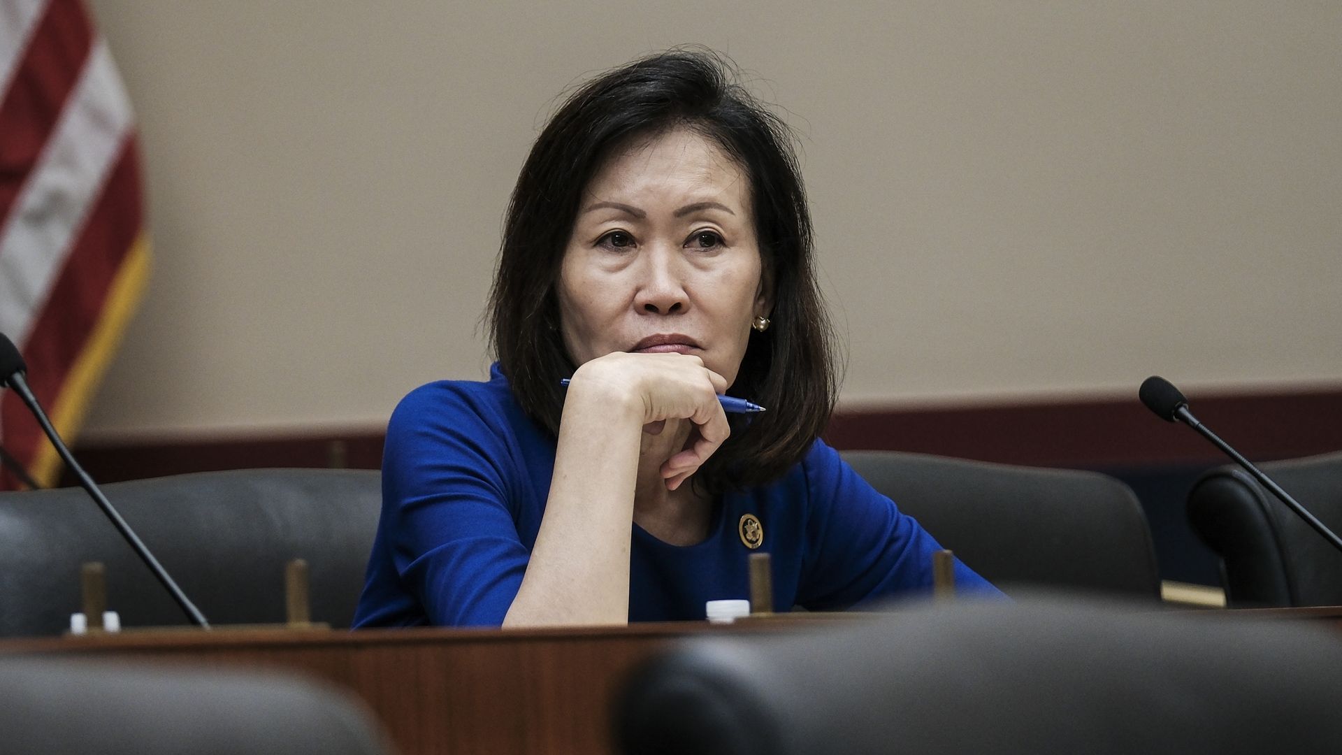 Rep. Michelle Steel, wearing a blue dress and sitting at a podium with her hand holding a pen in front of her chin.