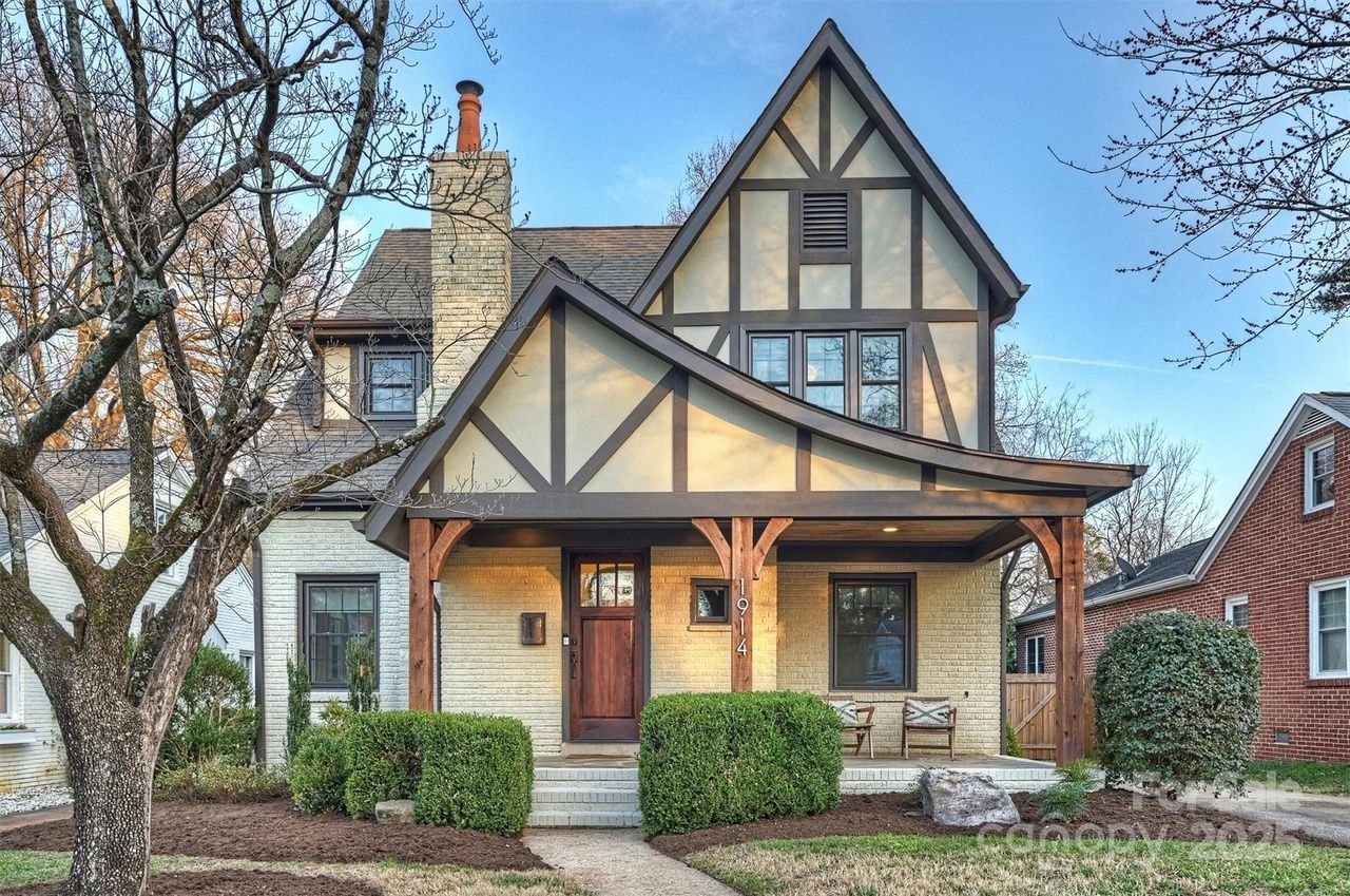 Two-story Tudor-style house with beige brick and dark wood trim, curved porch roof with wooden beams, brown front door, and leafless tree in front under clear blue sky.