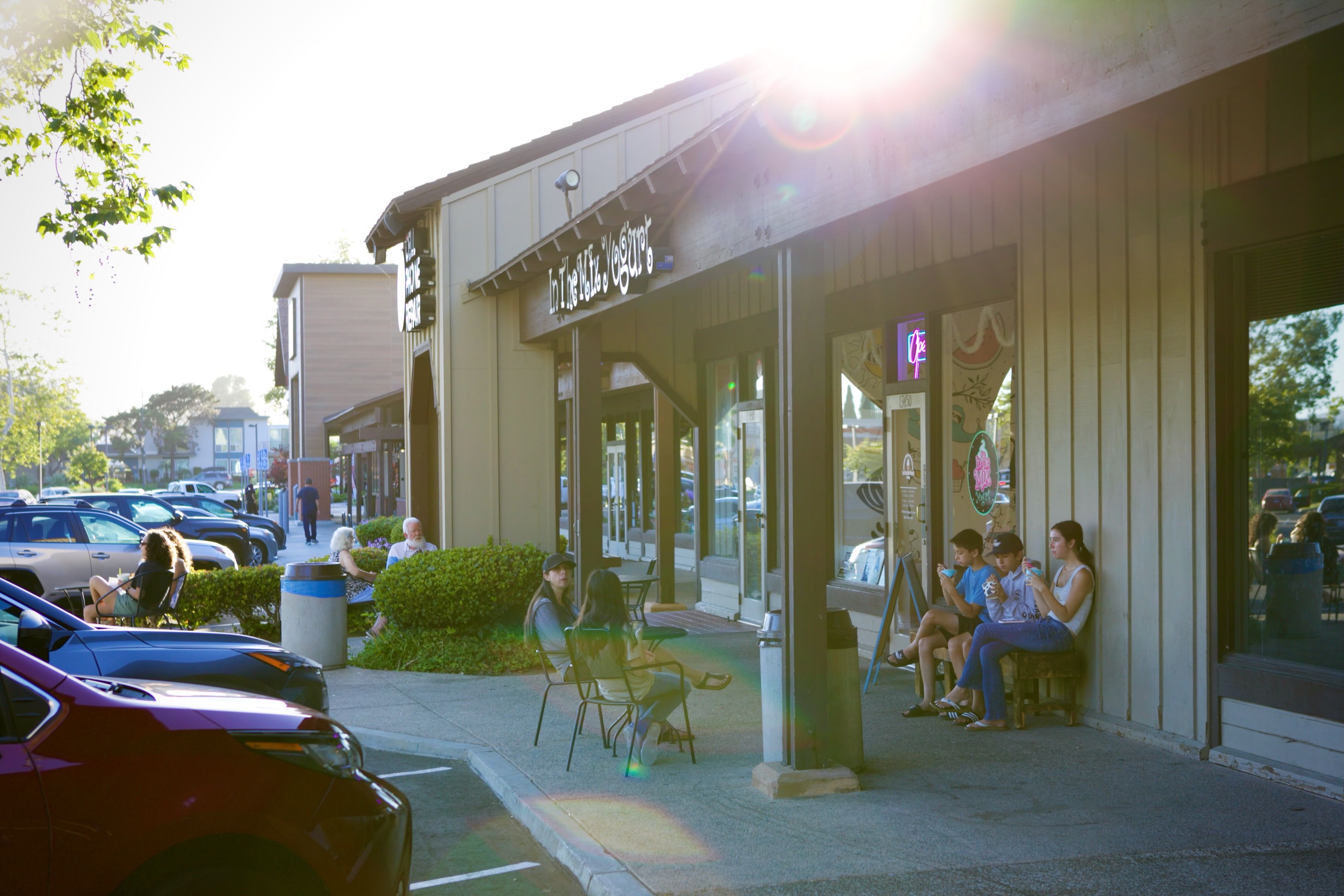 Sunny street scene showing a row of storefronts, people sitting at outdoor tables, parked cars along the curb, and leafy trees on the left; bright sun flare over the building.