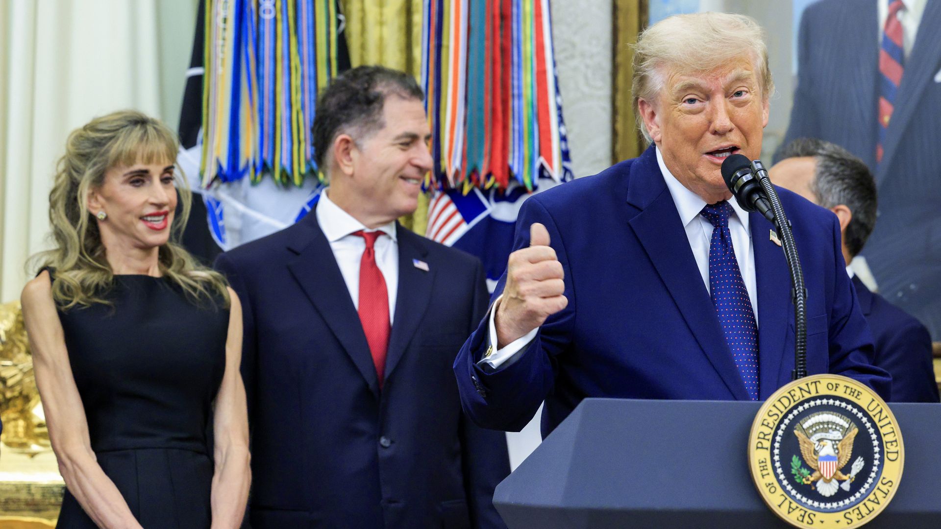Donald Trump speaks at a podium with the presidential seal, wearing a dark blue suit and dotted tie, with Michael and Susan Dell behind him; colorful ribbons behind.