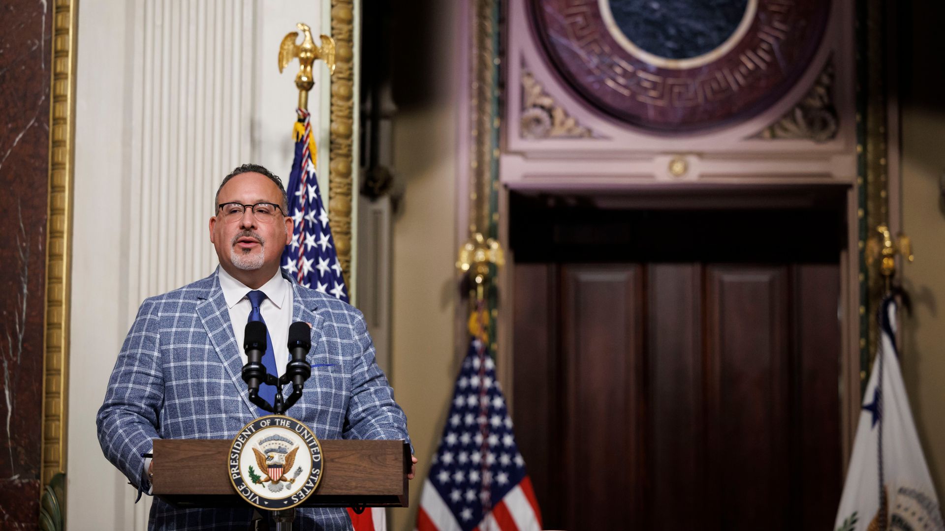 Miguel Cardona speaks at a lectern wearing a patterned suit with the American flag in the background 