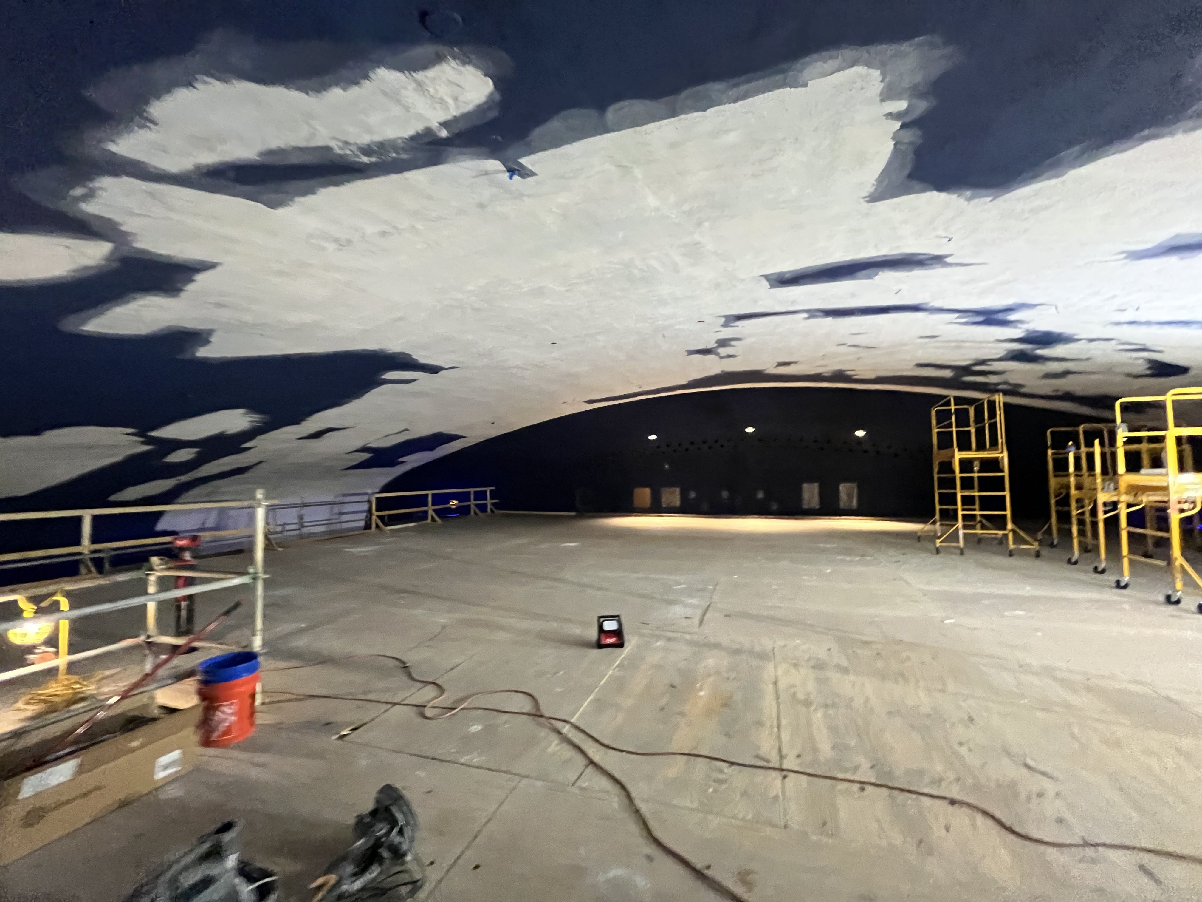 Inside a large indoor space with a dark blue ceiling showing white cloudlike patches. Yellow scaffolding on the right, railing along the left, and scattered cables, tools, and orange buckets on the concrete floor.
