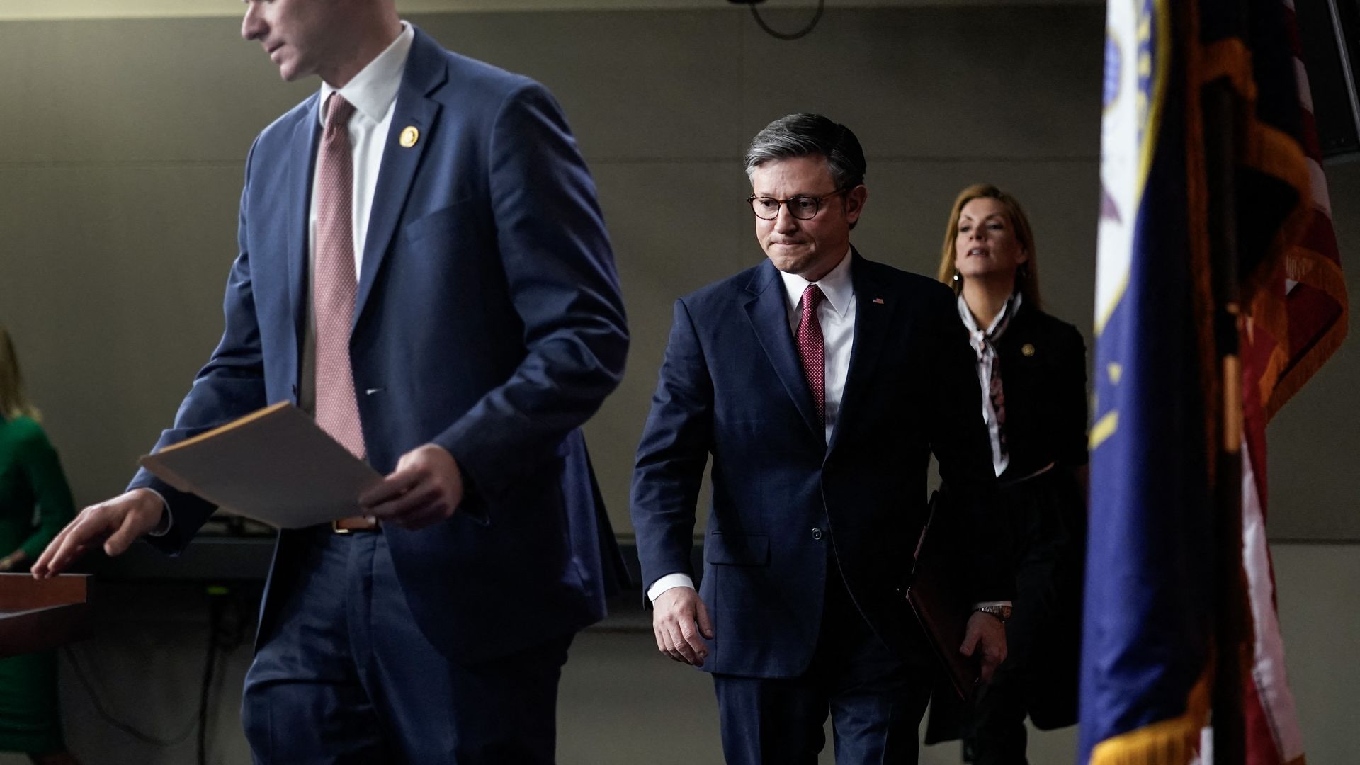 House Speaker Mike Johnson, wearing a blue suit, white shirt, red tie and glasses, walks up to the podium in Studio A in the U.S. Capitol.