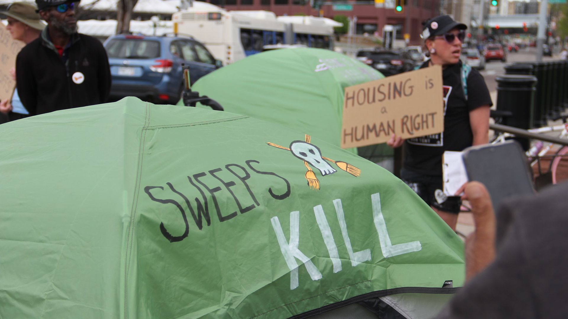 A light green camping tent with the word SWEEPS KILL and a white skull is seen next to two people, including one person holding up a cardboard sign with the words HOUSING IS A HUMAN RIGHT. In the foreground, a man holds up his cellphone. 