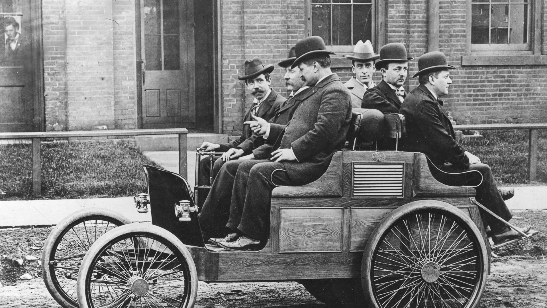 Black-and-white photo of men in bowler hats and suits riding in an open wooden carriage with large wheels, parked in front of a brick building; sign above reads "The Ninton Motor Carriage Co."