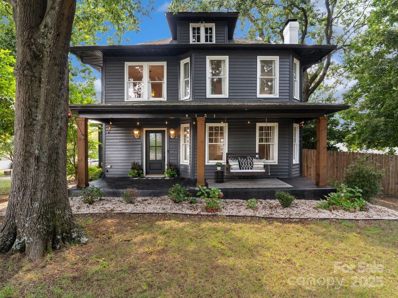 Two-story dark gray house with white-trimmed windows, a covered porch with wooden columns, a swing with cushions, and a large tree in front yard under blue sky.