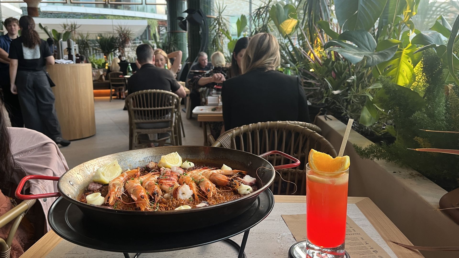 Outdoor restaurant scene with a large pan of seafood paella in the foreground, lemon wedges, and a bright red-orange cocktail on the table. Diners sit on wicker chairs among lush green plants as staff move in the background.