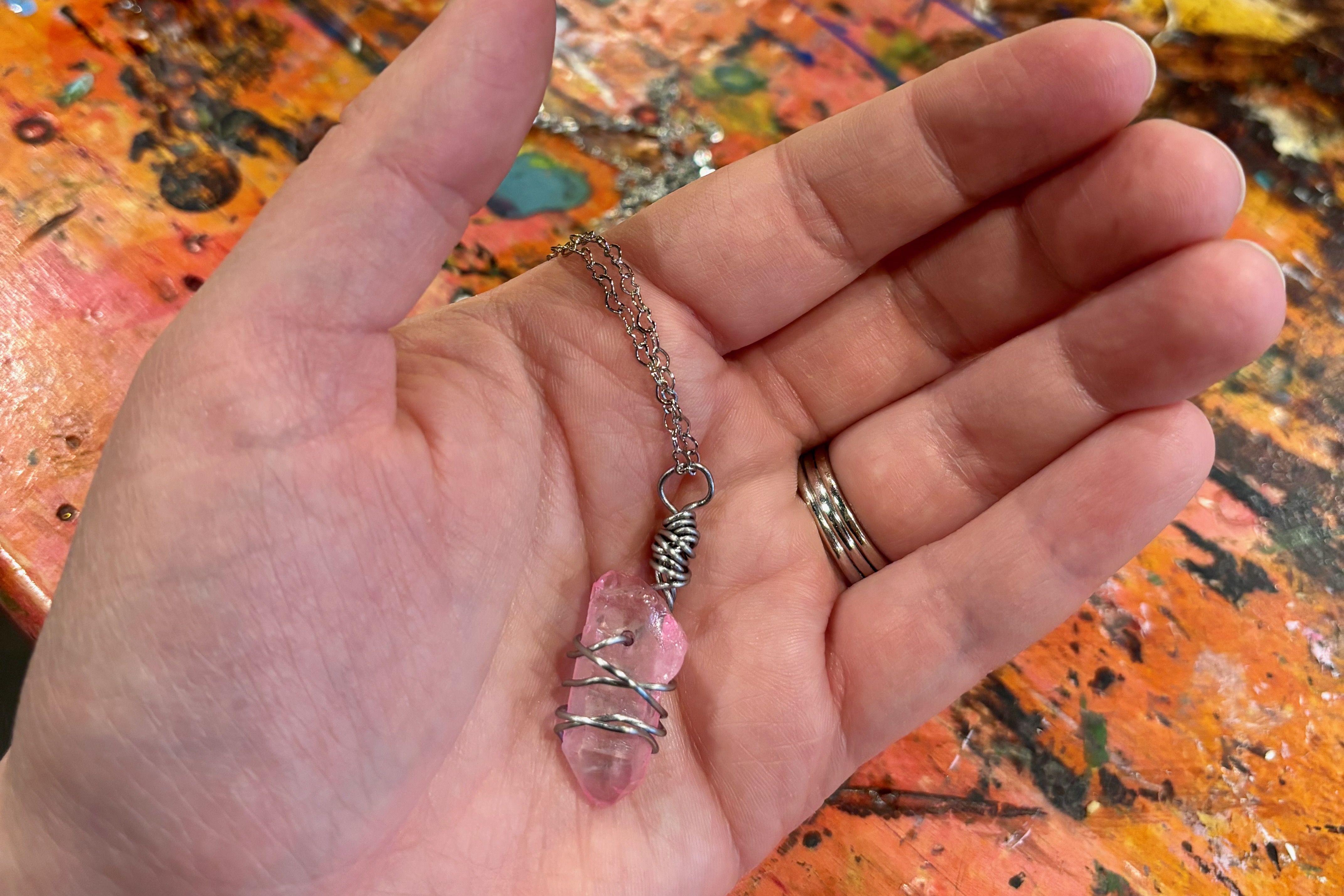 Hand holding a silver chain necklace with a pink crystal wrapped in silver wire, against a colorful, paint-splattered wooden surface background.