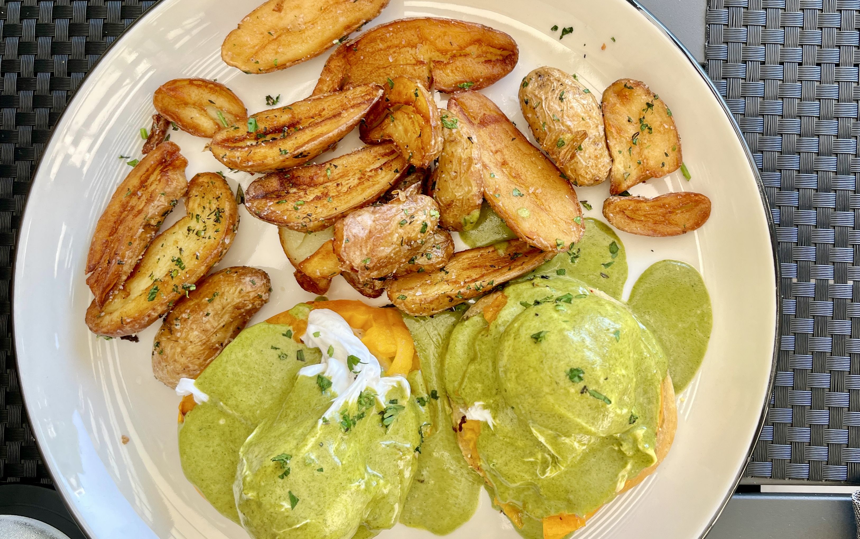 White plate with roasted potato wedges and eggs Benedict topped with green sauce and herbs, on a woven black and gray placemat.