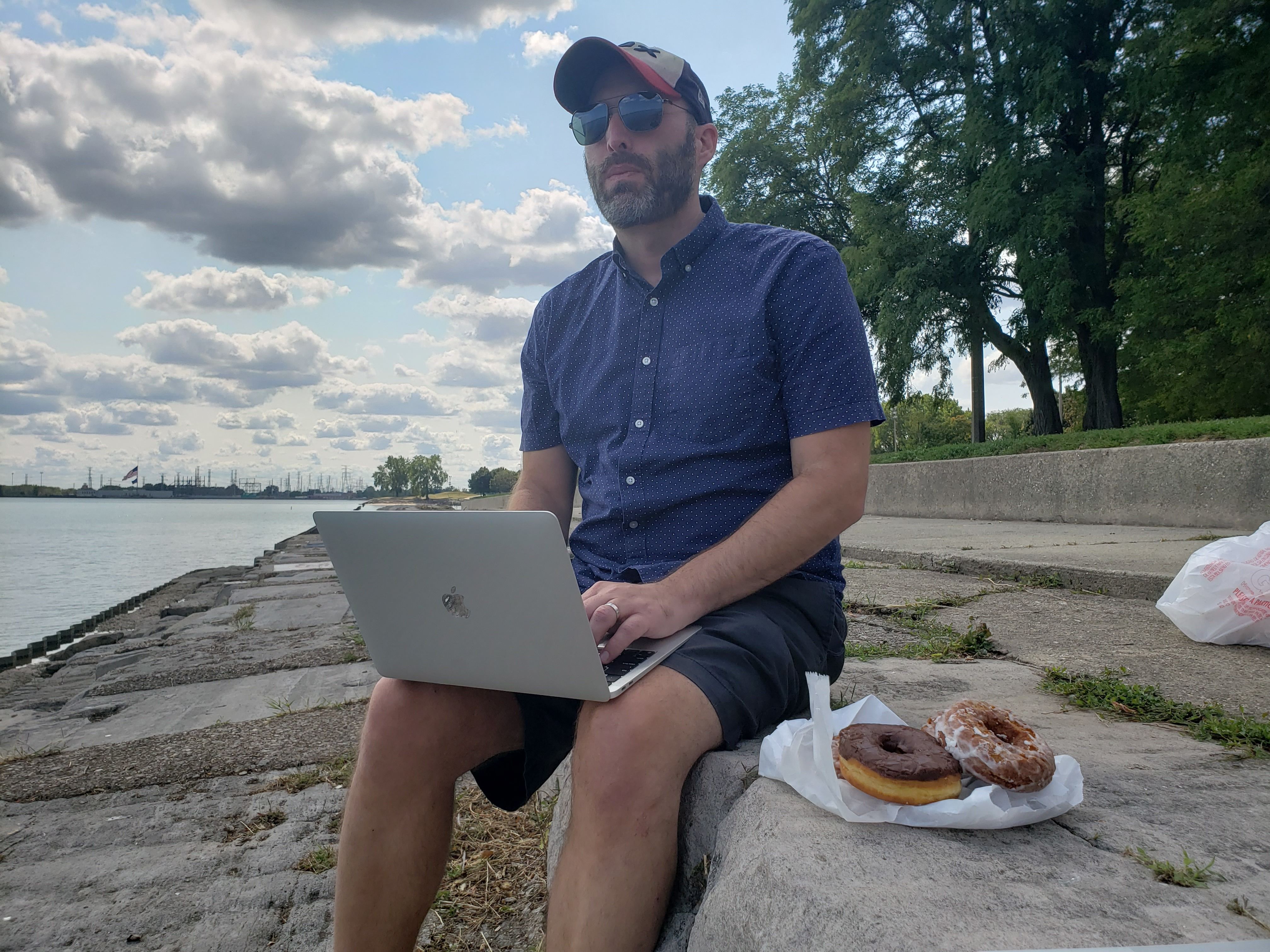 Photo of a man sitting on a rock with a computer in his hands and doughnuts next to him. 
