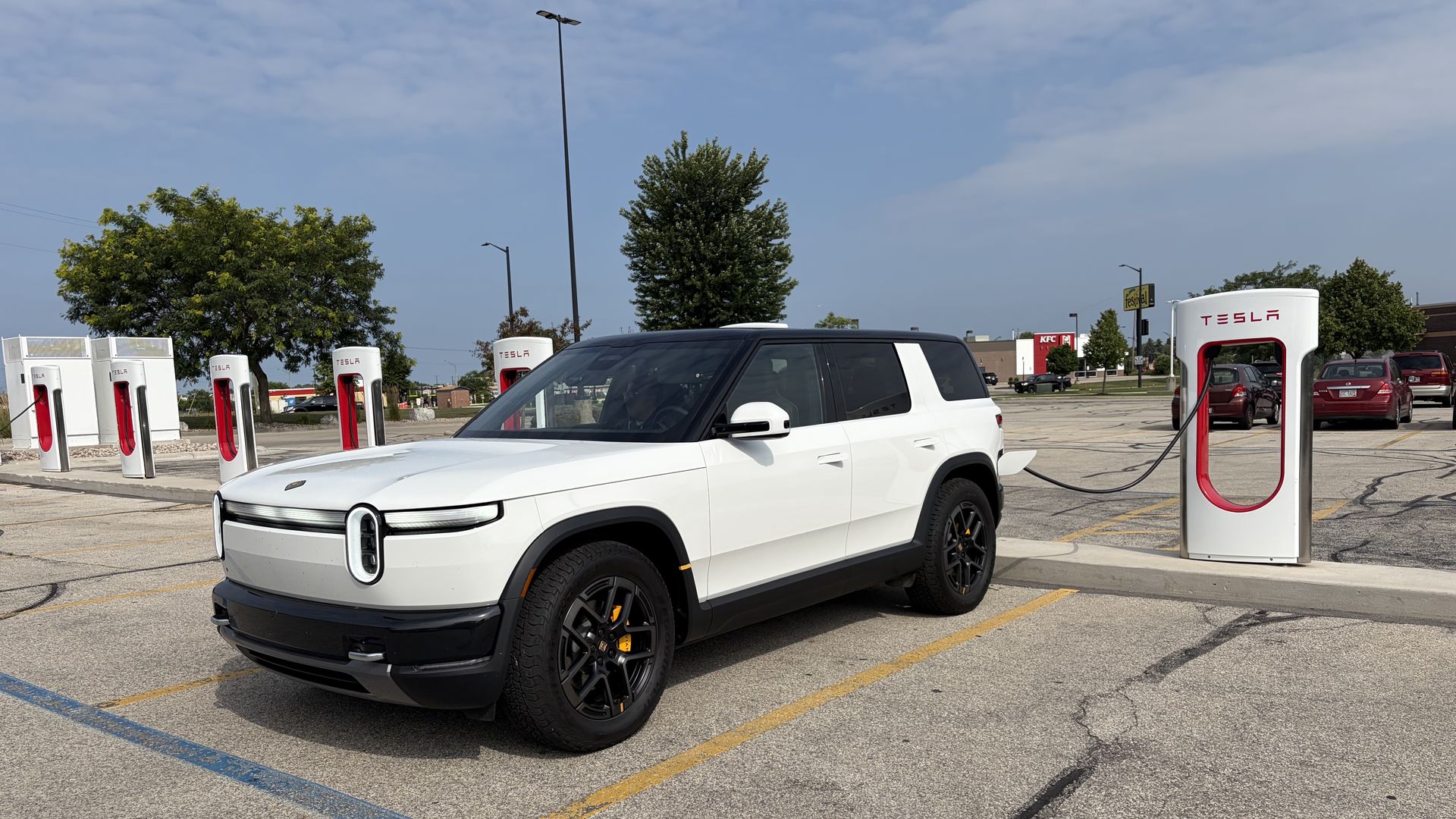 White Rivian R2 SUV plugged into a Tesla Supercharger station in a parking lot. Several white TESLA charging kiosks line the foreground; blue sky, trees, and distant buildings with a KFC sign.