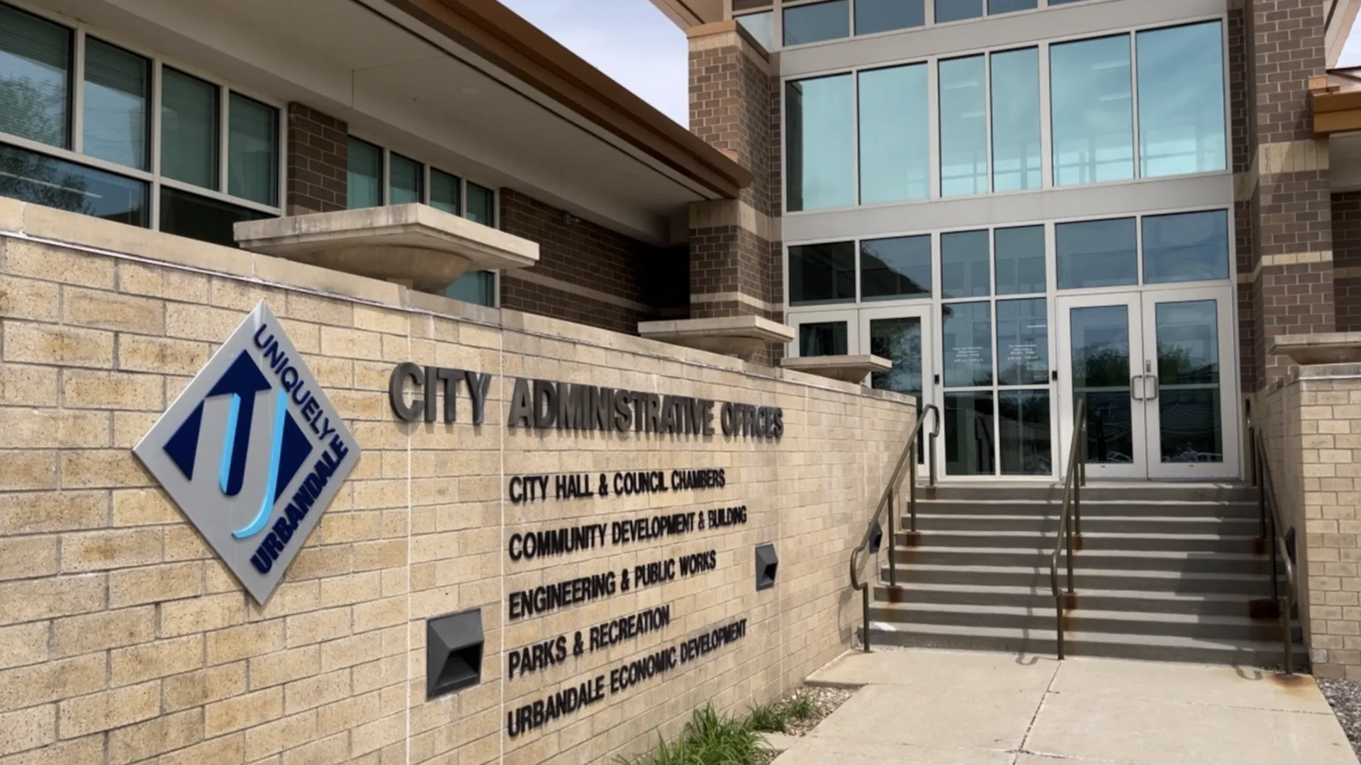 Exterior of a municipal building with beige brick and a glass entry. A wall sign reads "CITY ADMINISTRATIVE OFFICES" and a blue diamond sign says "Uniquely Urbana" beside the stairs.