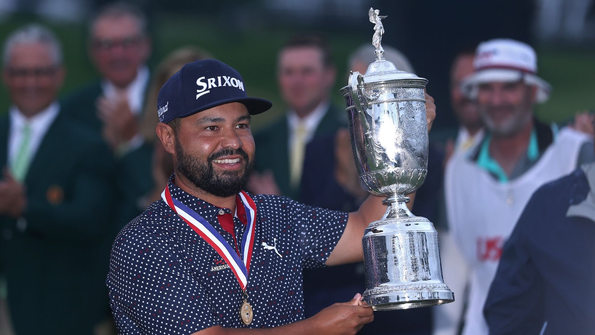 With a medal around his neck, golfer J.J. Spaun holds up a trophy after winning the 2025 U.S. Open.