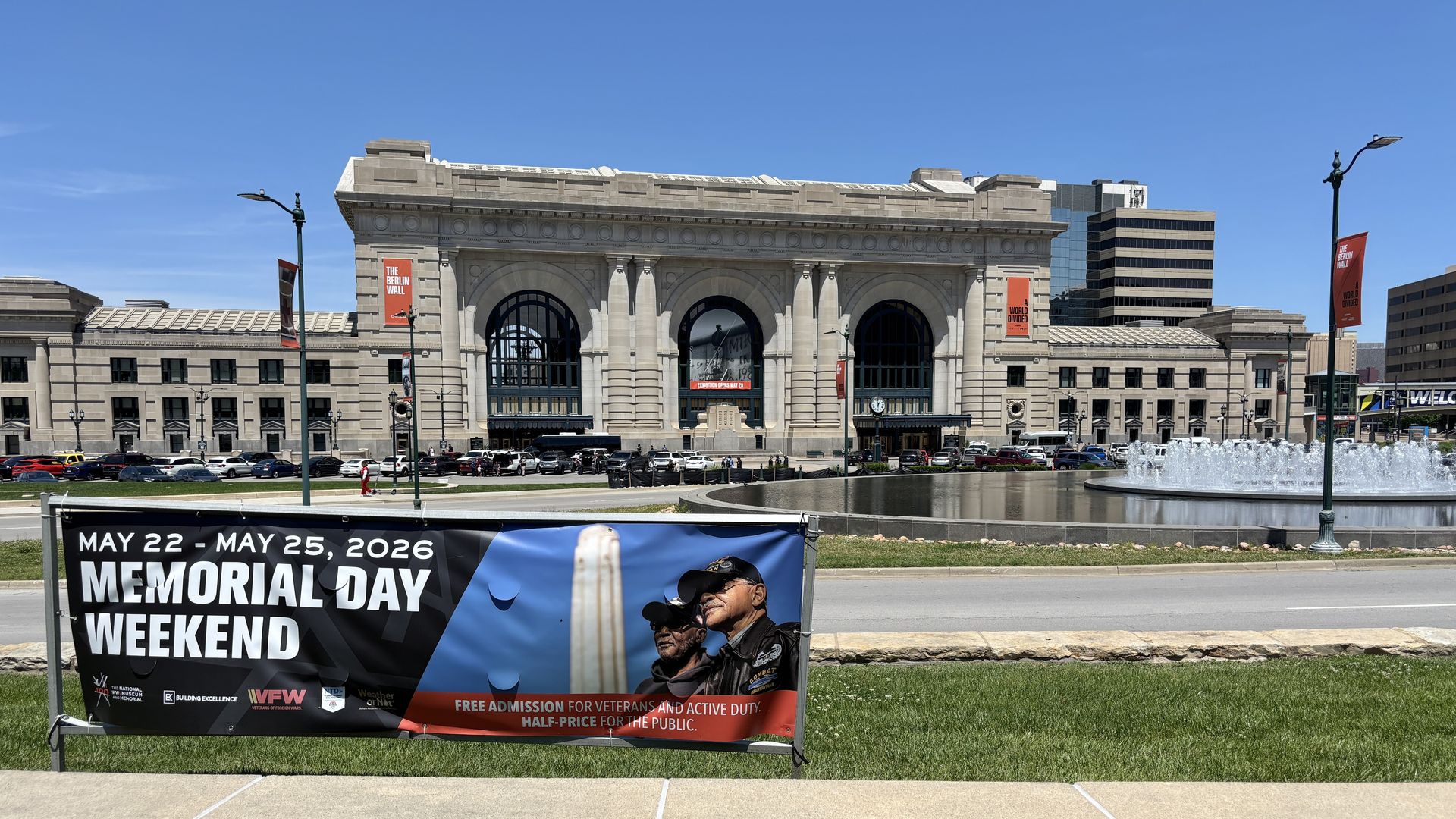 Grand stone building with three arched windows and columns under a blue sky. Red banners read "The Berlin Wall". A Memorial Day Weekend banner (May 22–25, 2026) shows veterans; fountain and cars line the street.