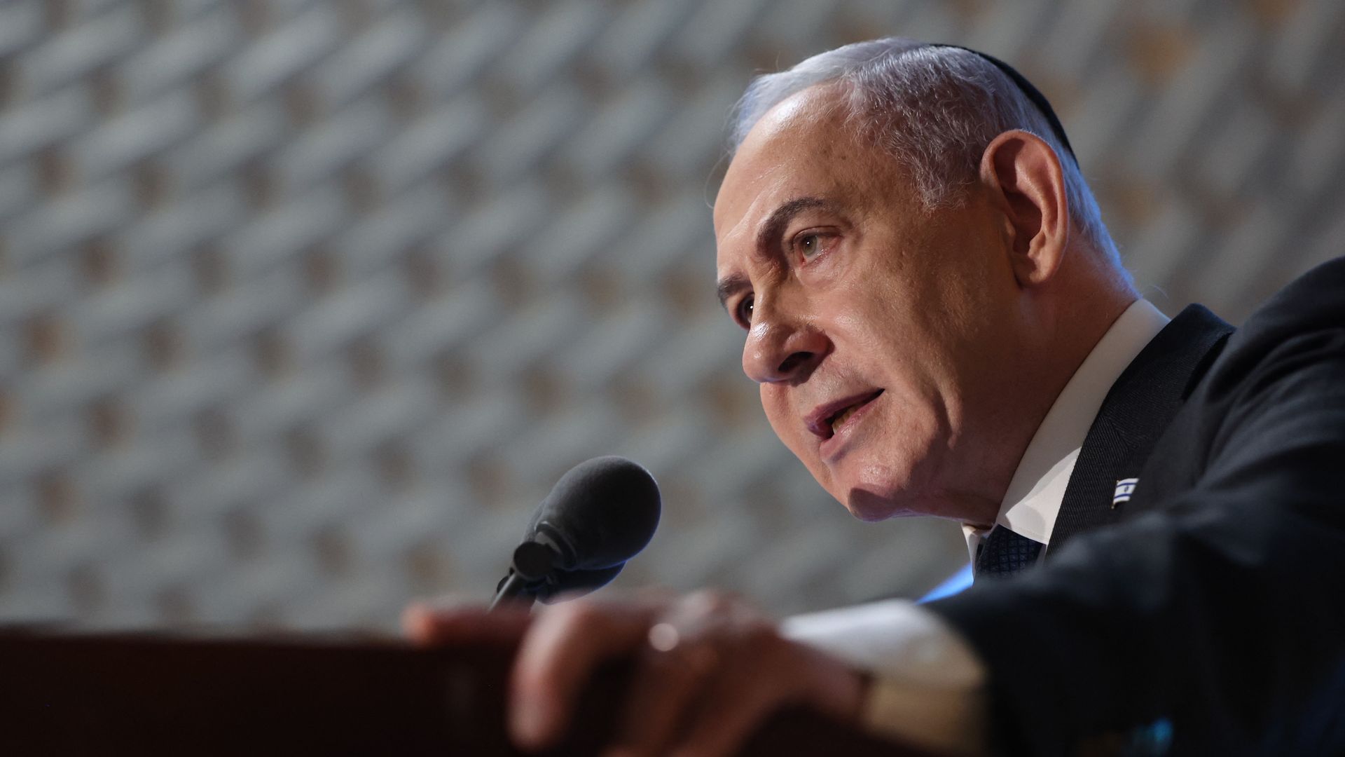 Benjamin Netanyahu, wearing a gray suit and a yarmulke and speaking into a microphone below a white brick ceiling.