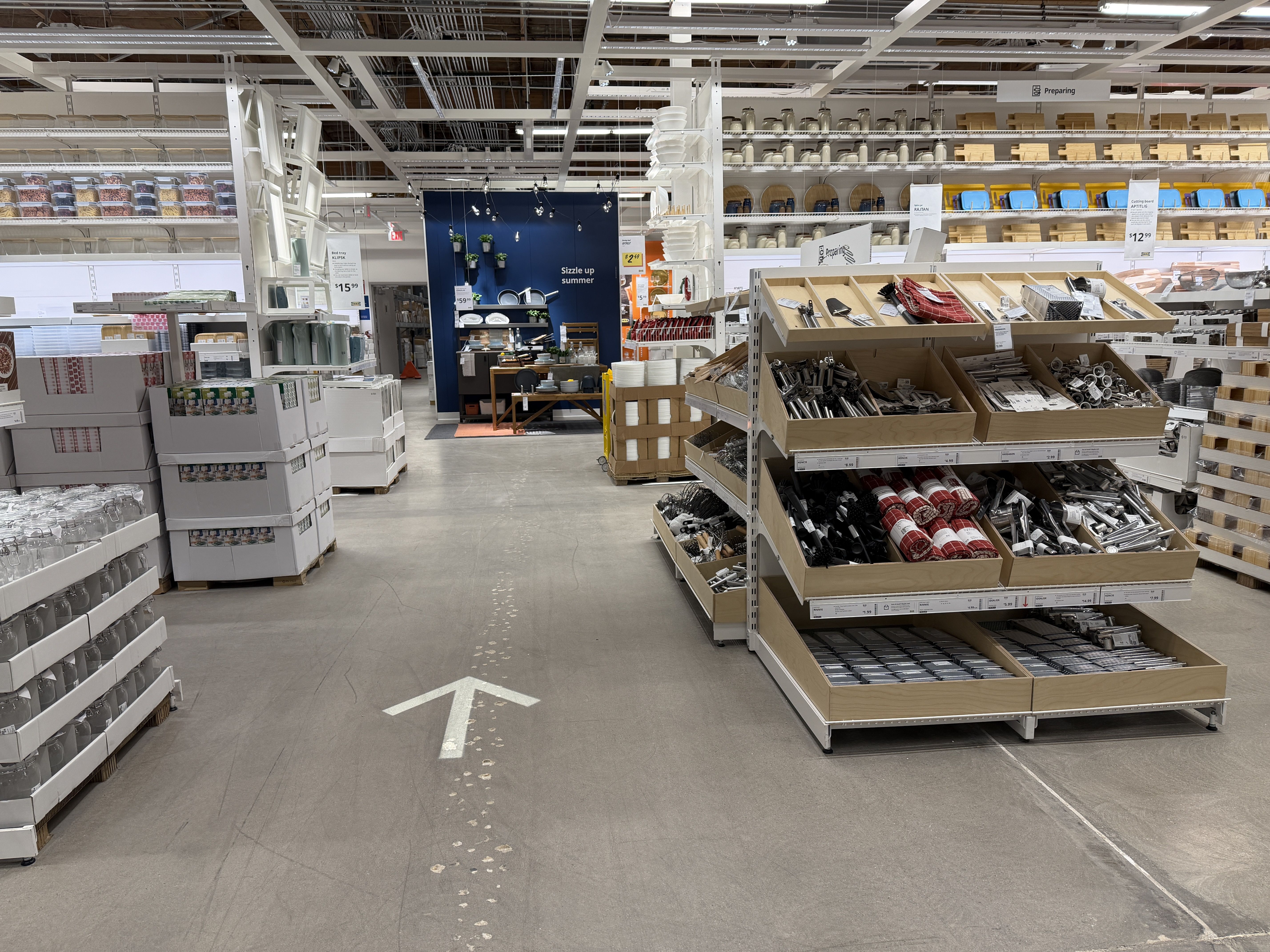 Spacious home goods store with an industrial ceiling. A central aisle features metal and wood display shelves stocked with kitchenware and hardware; a white floor arrow points ahead toward the back area.