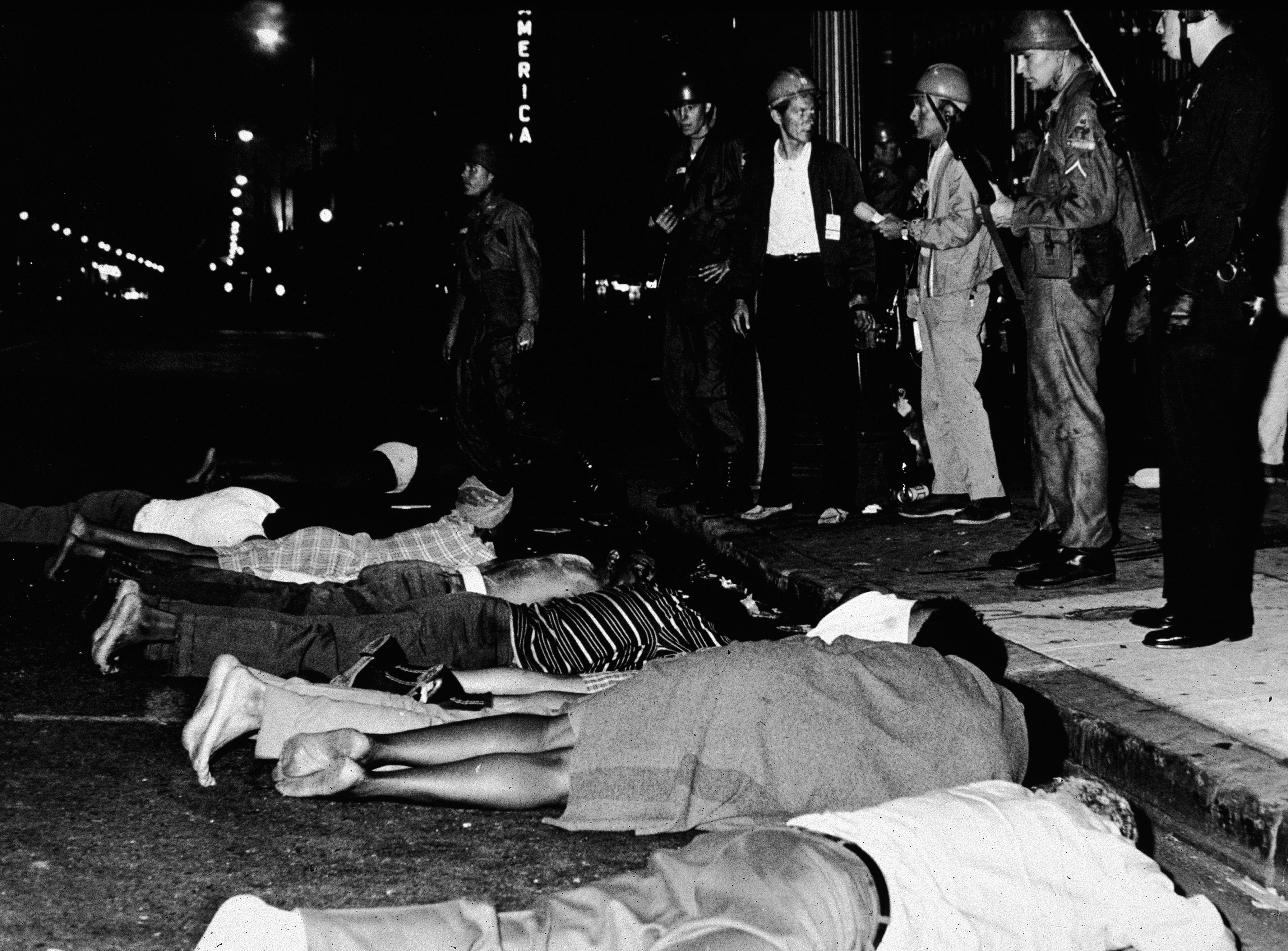 Black and white photo of people lying face down on a street at night, surrounded by uniformed soldiers and police officers standing on the sidewalk under streetlights.