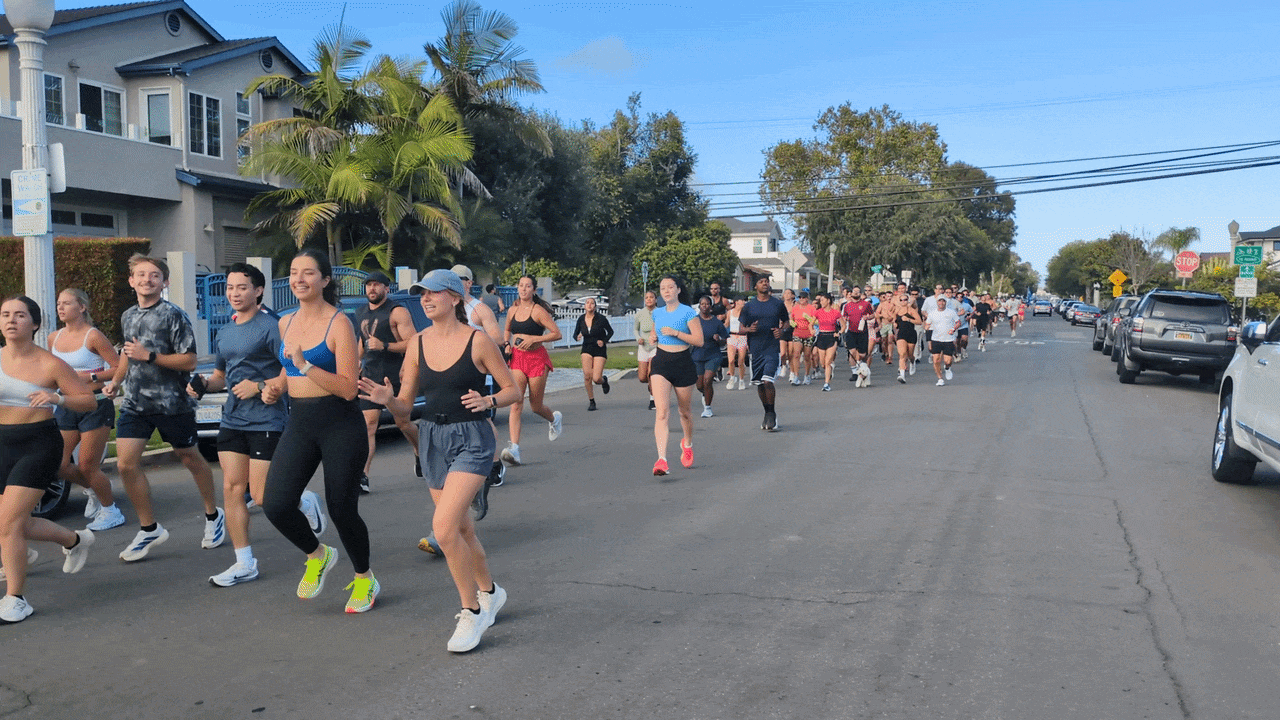 Runners participate in a sunny street race along a residential road, wearing colorful athletic gear. Palm trees line the curb as parked cars and a stop sign appear ahead.
