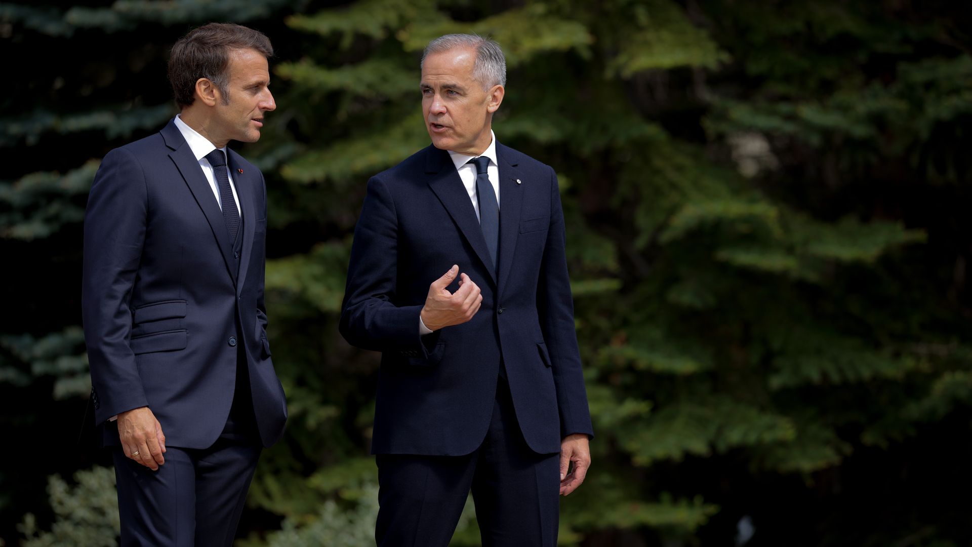 Two men in dark suits walking and talking outdoors, with green leafy trees in the background, one gesturing with hand while speaking.
