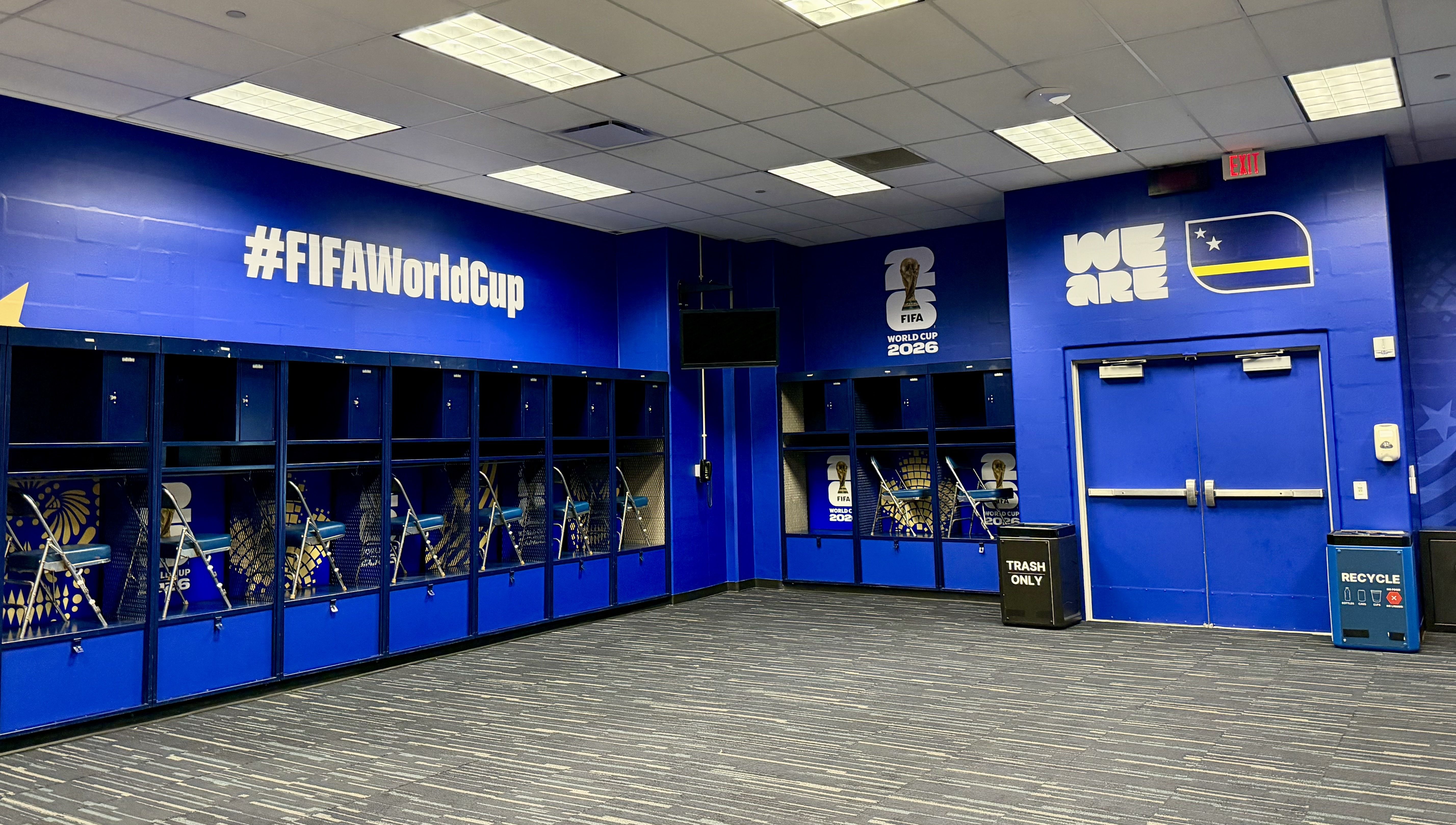 Blue locker room with a long row of lockers, chairs, and benches. Large white #FIFAWorldCup text; FIFA World Cup 2026 logo; we are with a flag on the wall; exit sign, trash and recycle bins.