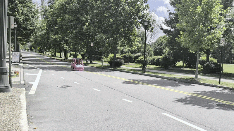 A gif showing a Power Wheels Barbie Camper Van go-kart driving quickly on a tree-lined street.
