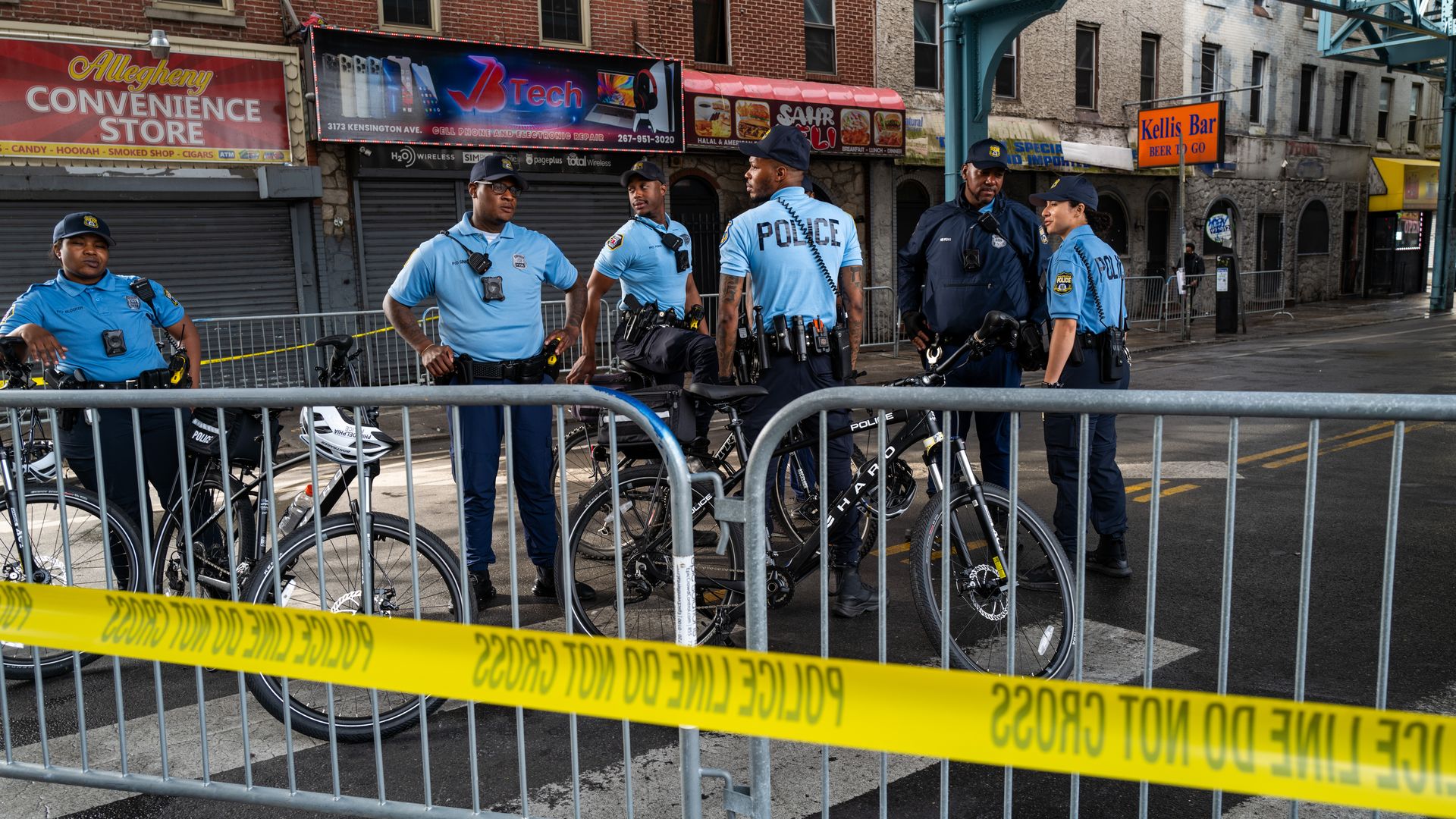 Several Philadelphia officers stand near bikes in front of barrier fencing and police tape.