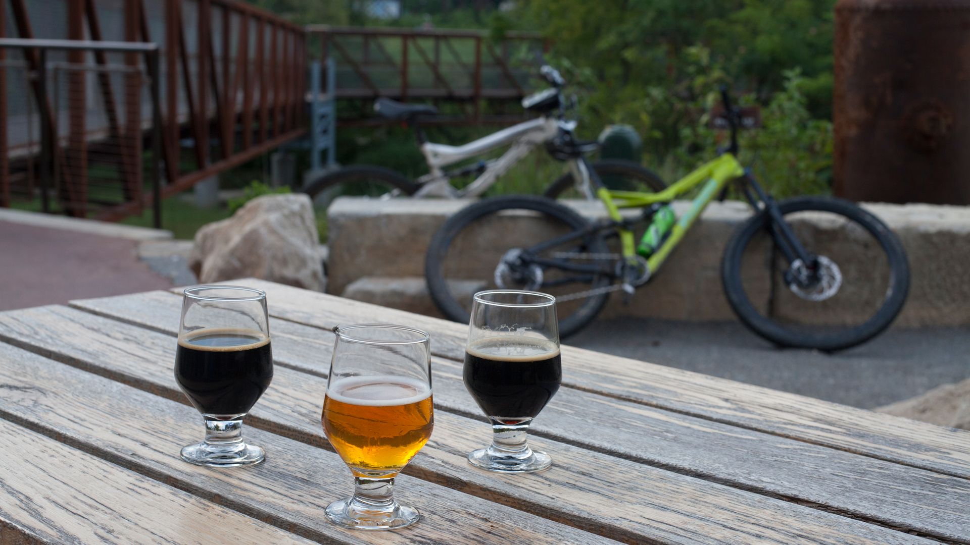 Four beer glasses on a rustic wooden table outdoors, with three dark beers and one amber in the center, while blurred bicycles and green foliage fill the background.