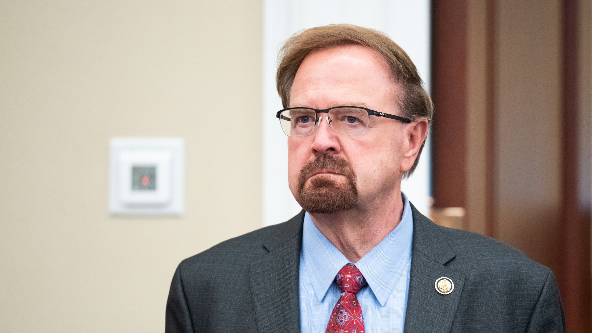 Close-up of a middle-aged man with light brown hair, goatee, and glasses, in a dark suit, light blue shirt, and red patterned tie; beige wall and wood paneling visible behind him.