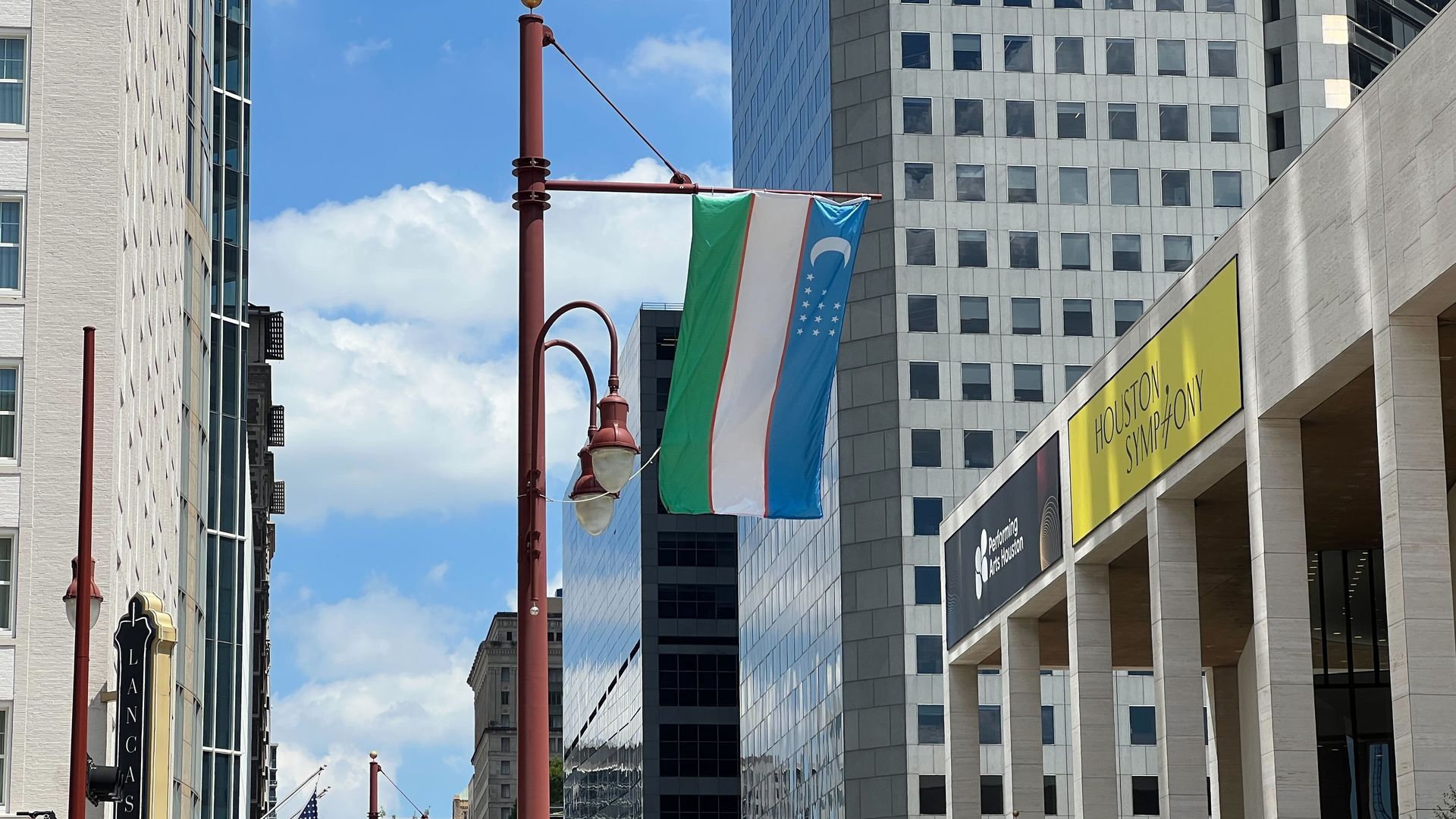 City street with tall modern buildings, red streetlights, and hanging flags (green, white, blue with crescent and stars). A yellow Houston Symphony banner on the right; American flag visible; blue sky.