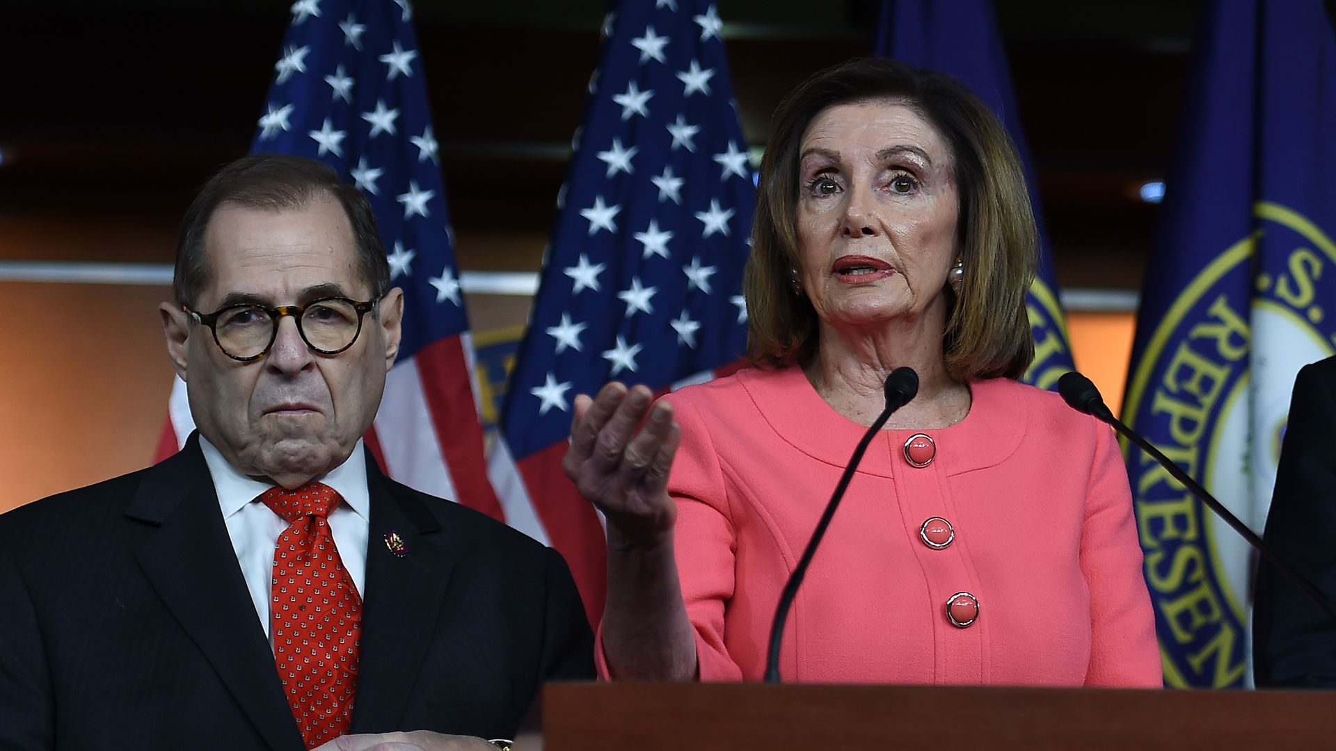 Rep. Jerry Nadler wearing a dark suit and then-Speaker Nancy Pelosi wearing a pink outfit standing in front of flags.