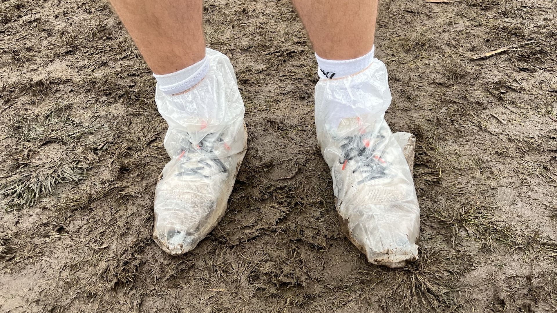 Two legs in white disposable boot covers stand on muddy, brown dirt ground with scattered twigs.