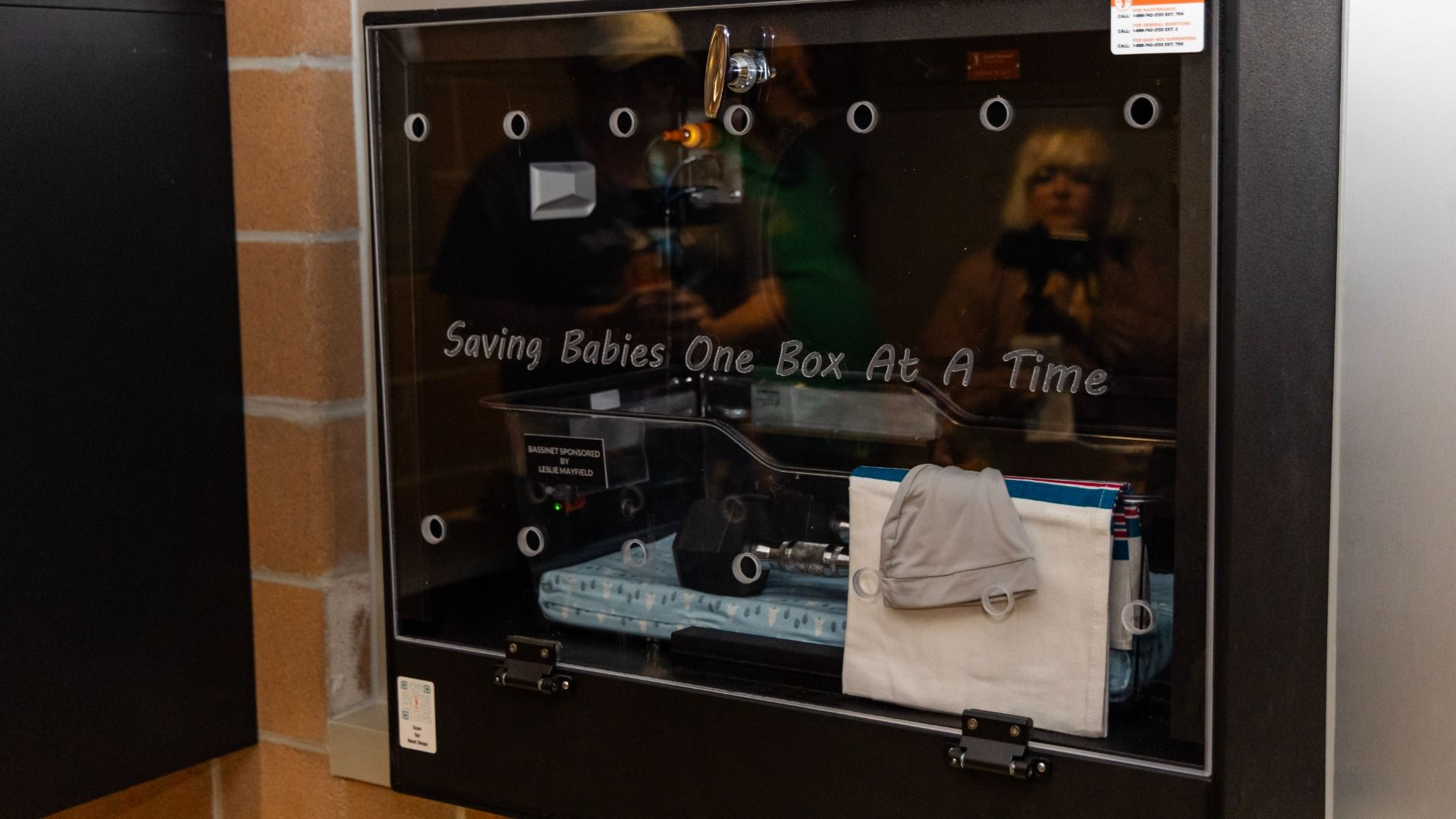 Black display cabinet with a glass front displaying "Saving Babies One Box At A Time." Inside: a blue-wrapped box, a gray bag, and a towel; reflections of people are seen in the glass.