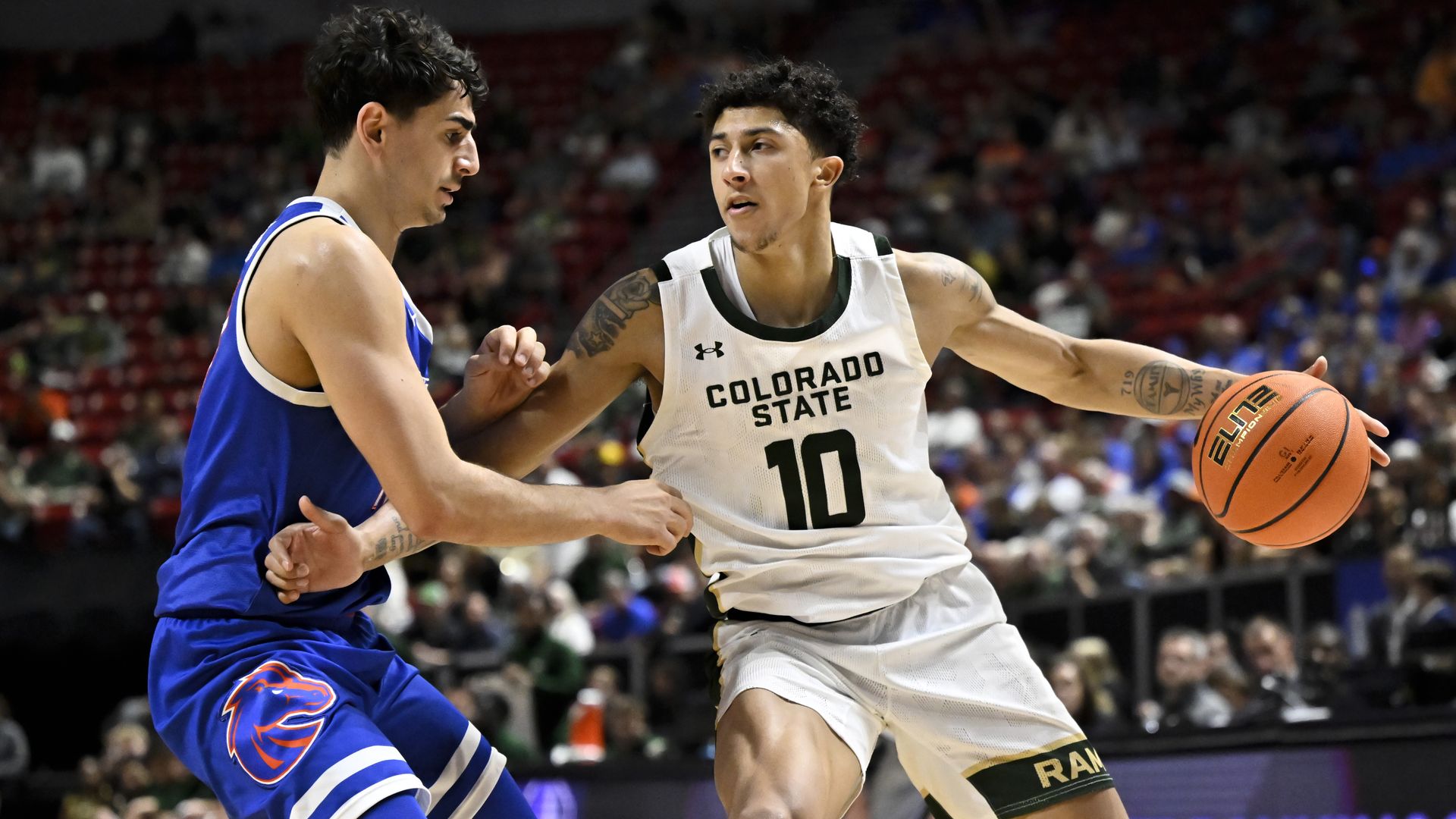 A basketball player in a white jersey with the words COLORADO STATE and the number 10 holds the ball while attempting to fend off another player. 