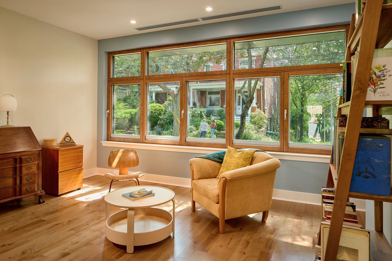 Cozy living room with large wooden-framed windows showing green trees and brick houses outside, featuring a brown armchair with pillows, a wooden chair, round white coffee table, and wooden furniture.