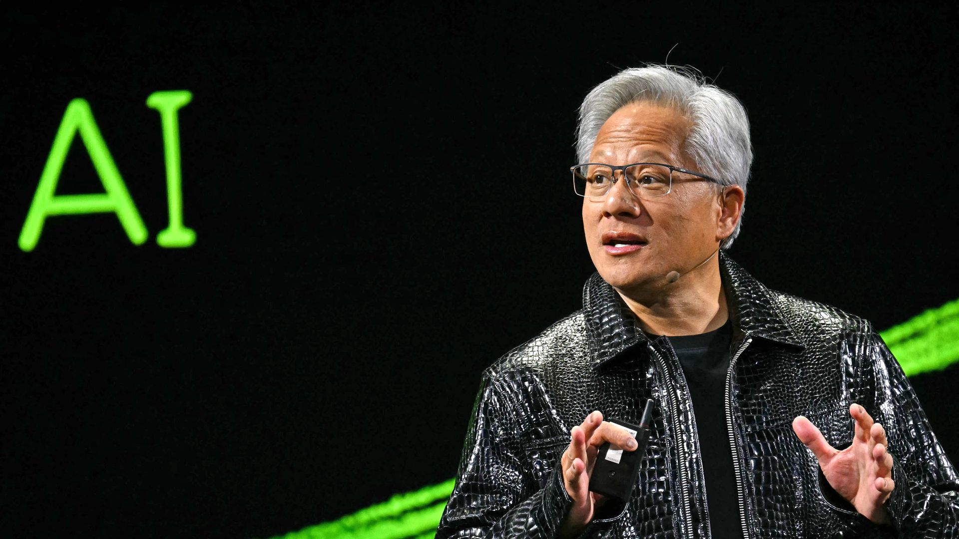 A man with gray hair and glasses speaks on stage, wearing a shiny black jacket with a headset mic; a bright green "AI" sign glows in the dark background.