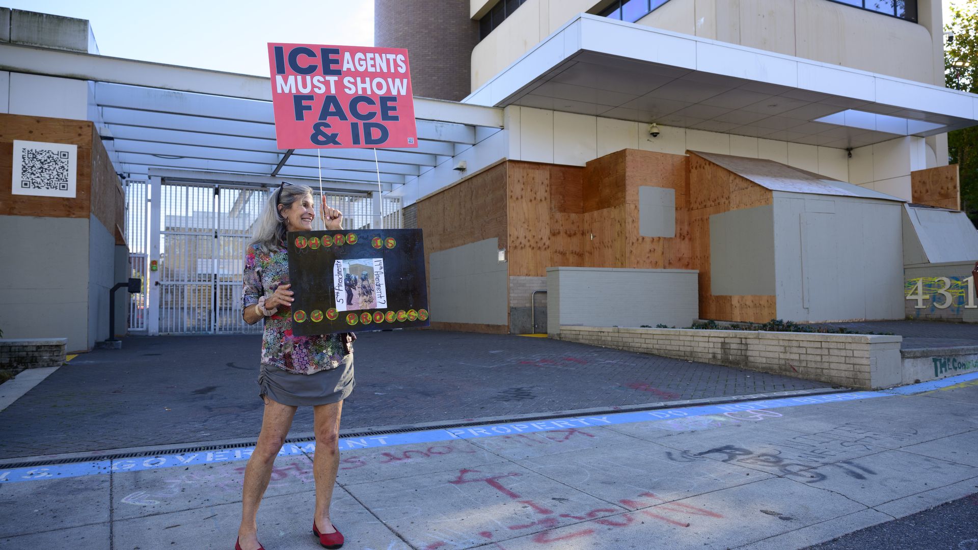 Woman in floral top and gray skirt stands outside a building with boarded-up windows holding two signs, one red saying "ICE agents must show face & ID" and another black with "Where is our process".