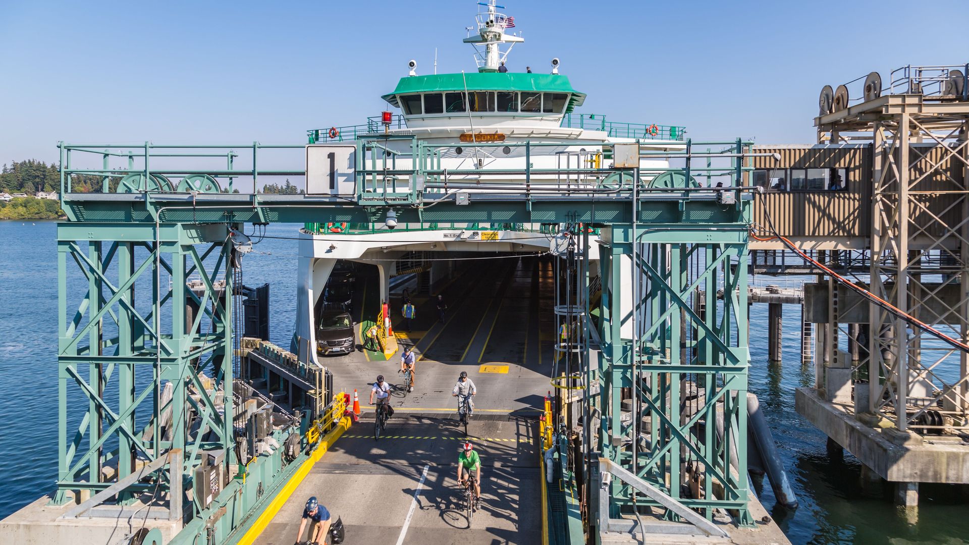 A white and sea-green colored ferry is seen docked as bicyclists exit onto a roadway. Light sea green scaffolding frames the roadway in front of the ferry.
