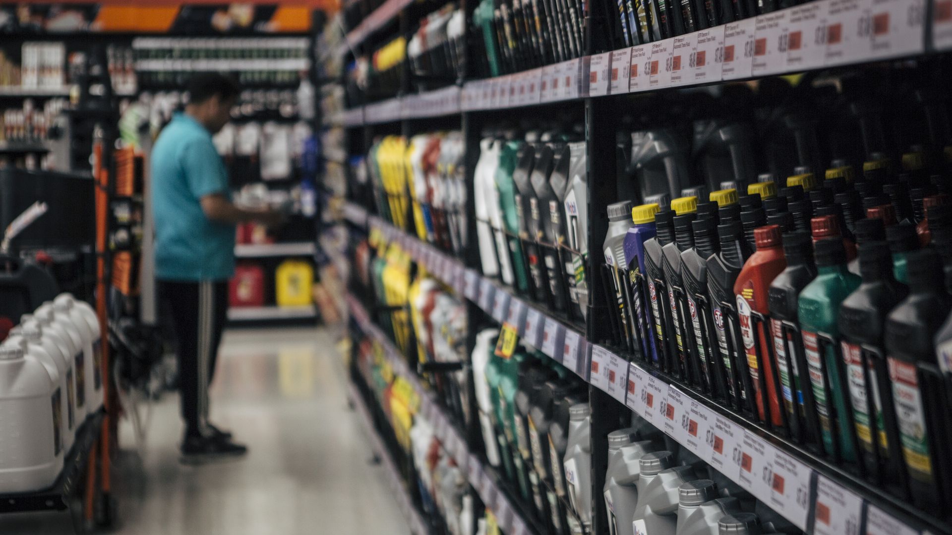 Blurred store aisle with shelves full of motor oil on the right: black bottles with yellow caps and price labels. A person in a teal shirt browses in the background.
