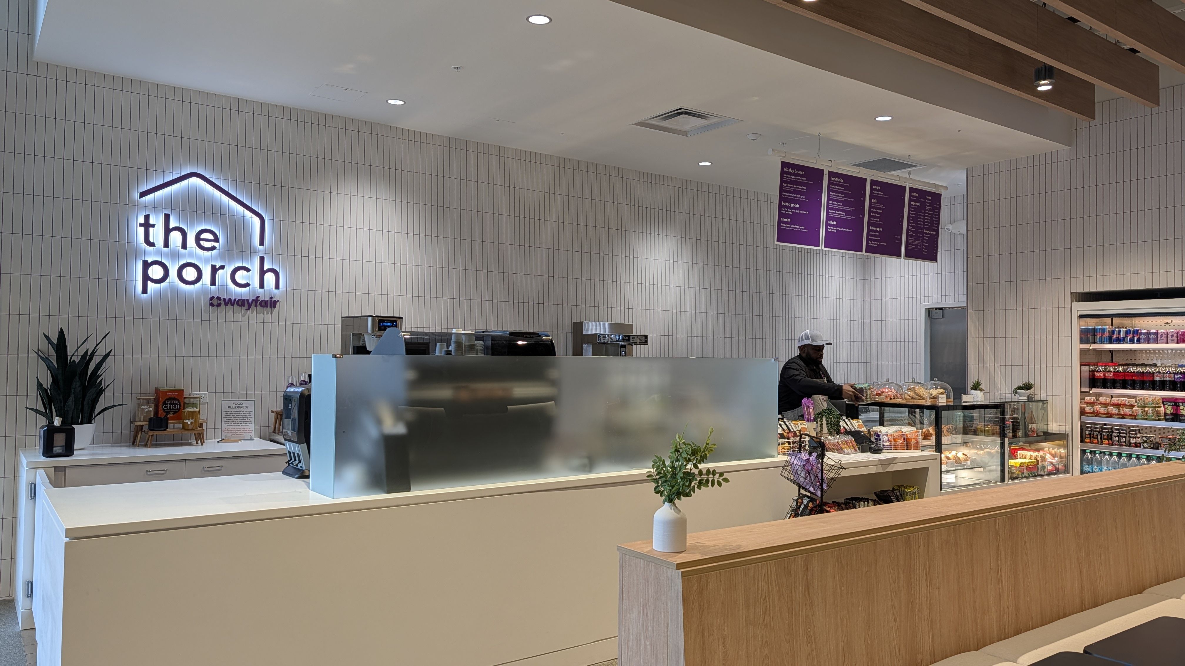 Interior view of a cafe called "the porch" with a neon sign on white tiled wall, white counter, frosted glass, pastry display, purple menu boards overhead, and a fridge to the right.