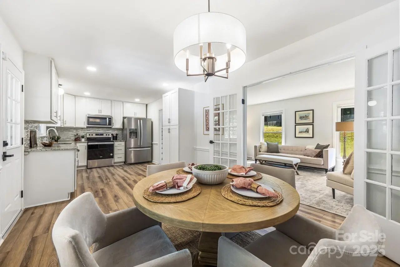 Bright open kitchen and dining area with white cabinets, stainless steel appliances, wood flooring, round wood table with place settings, beige upholstered chairs, and adjacent living room.