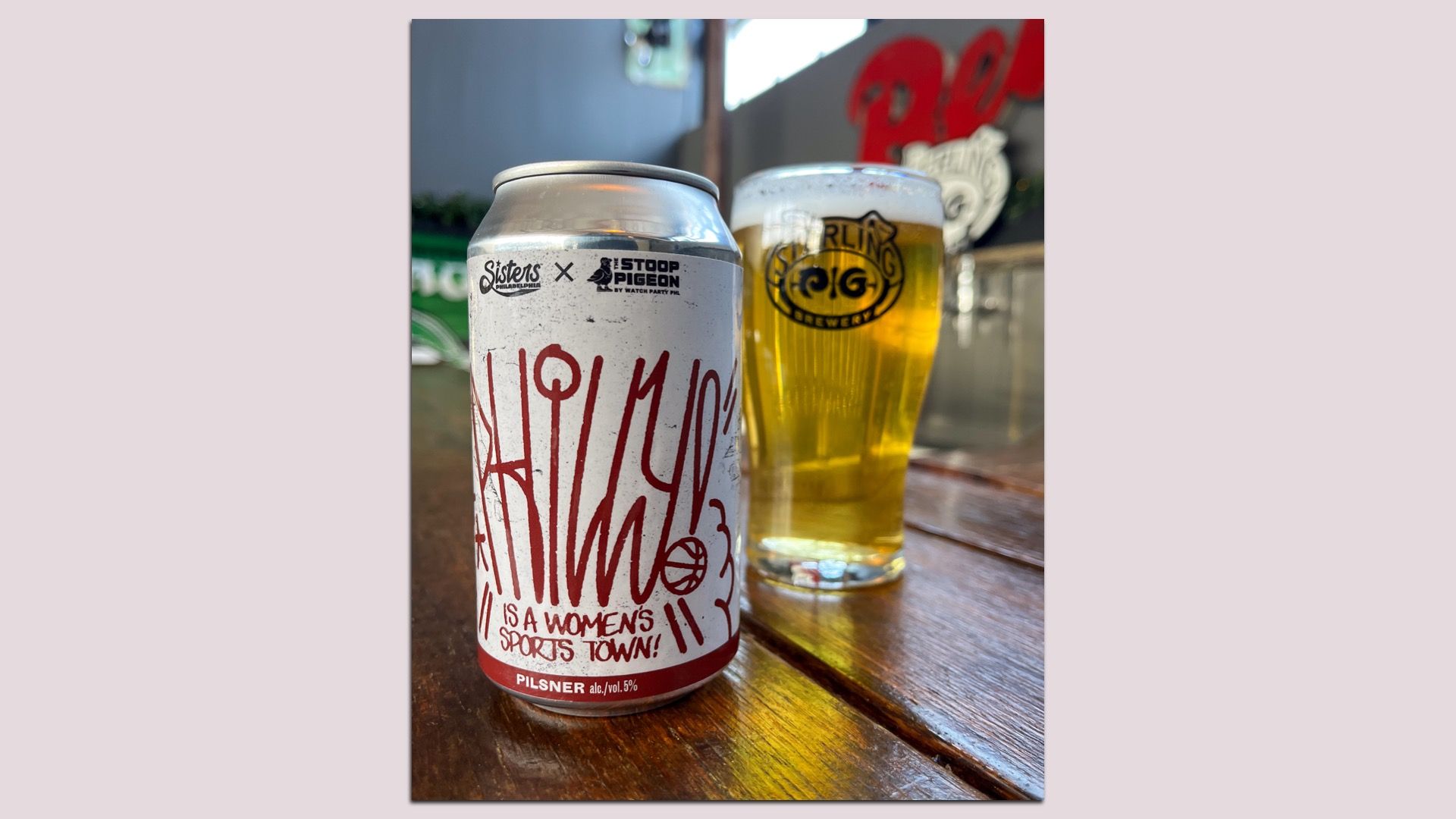 A can of pilsner with red graffiti-style text reading "This is a women's sports town!" next to a glass of golden beer with frothy head on a wooden table indoors.