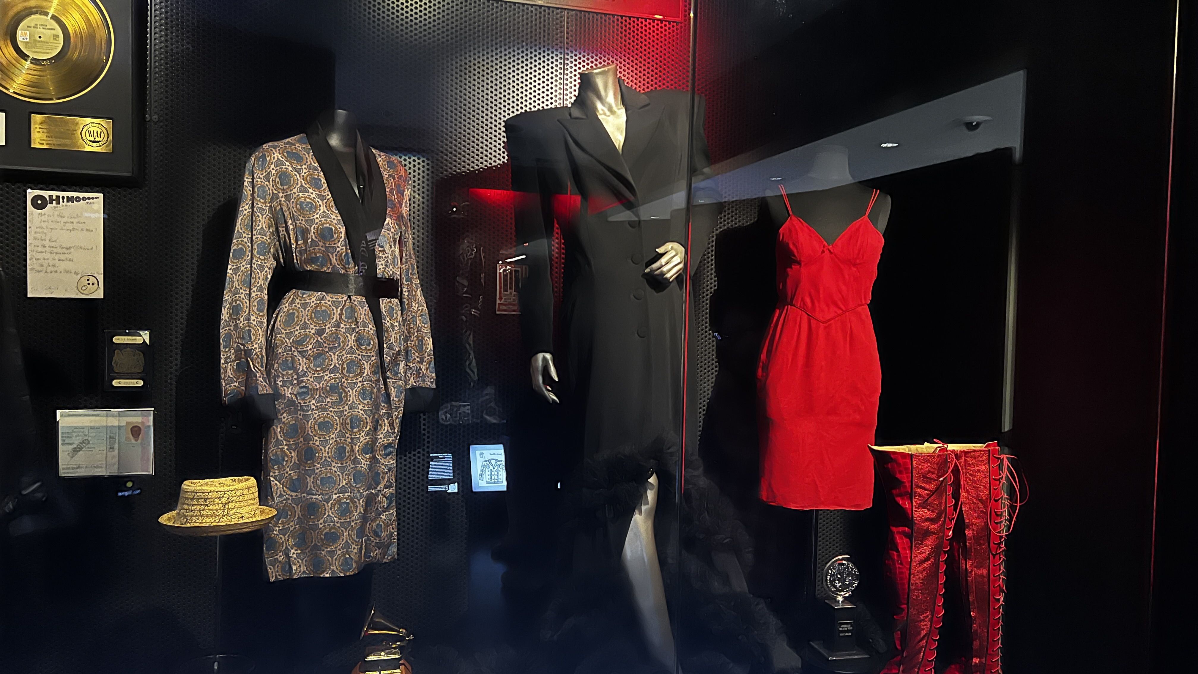 Display of three outfits in a dark room: a patterned robe with black belt and cuffs, a long black dress with ruffled hem, and a bright red dress with red thigh-high lace-up boots.