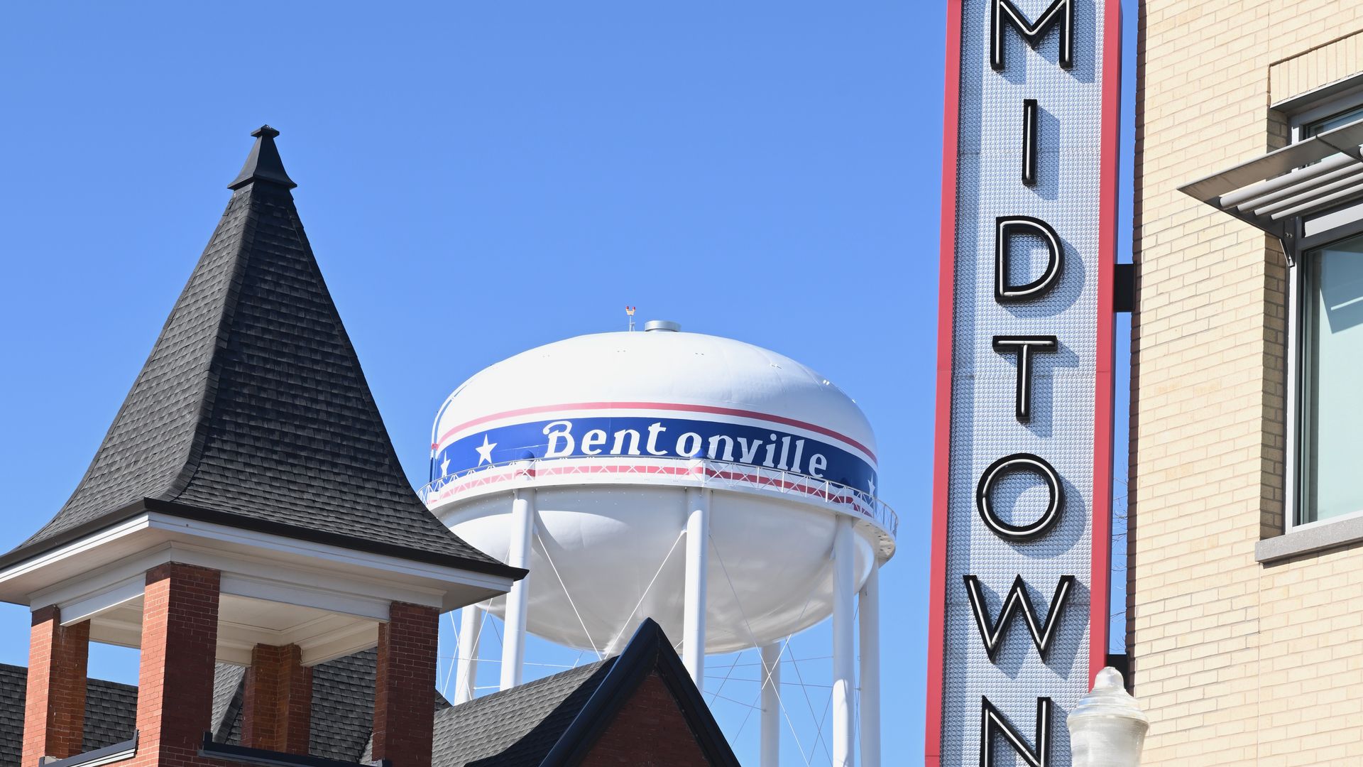 Water tower labeled "Bentonville" behind a brick building with a black spire and a vertical sign reading "MIDTOWN" against a clear blue sky.