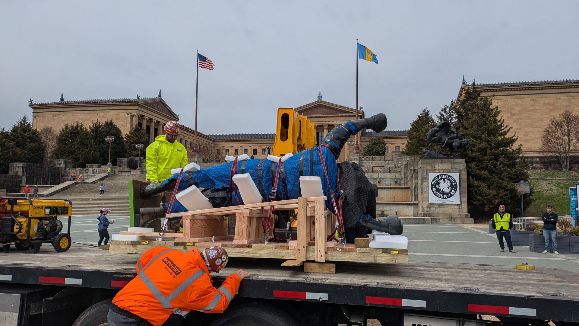 Workers in neon jackets unload a large blue-tarp sculpture on a wooden cradle from a flatbed trailer in front of the Philadelphia Museum of Art; US and Ukraine flags fly nearby.